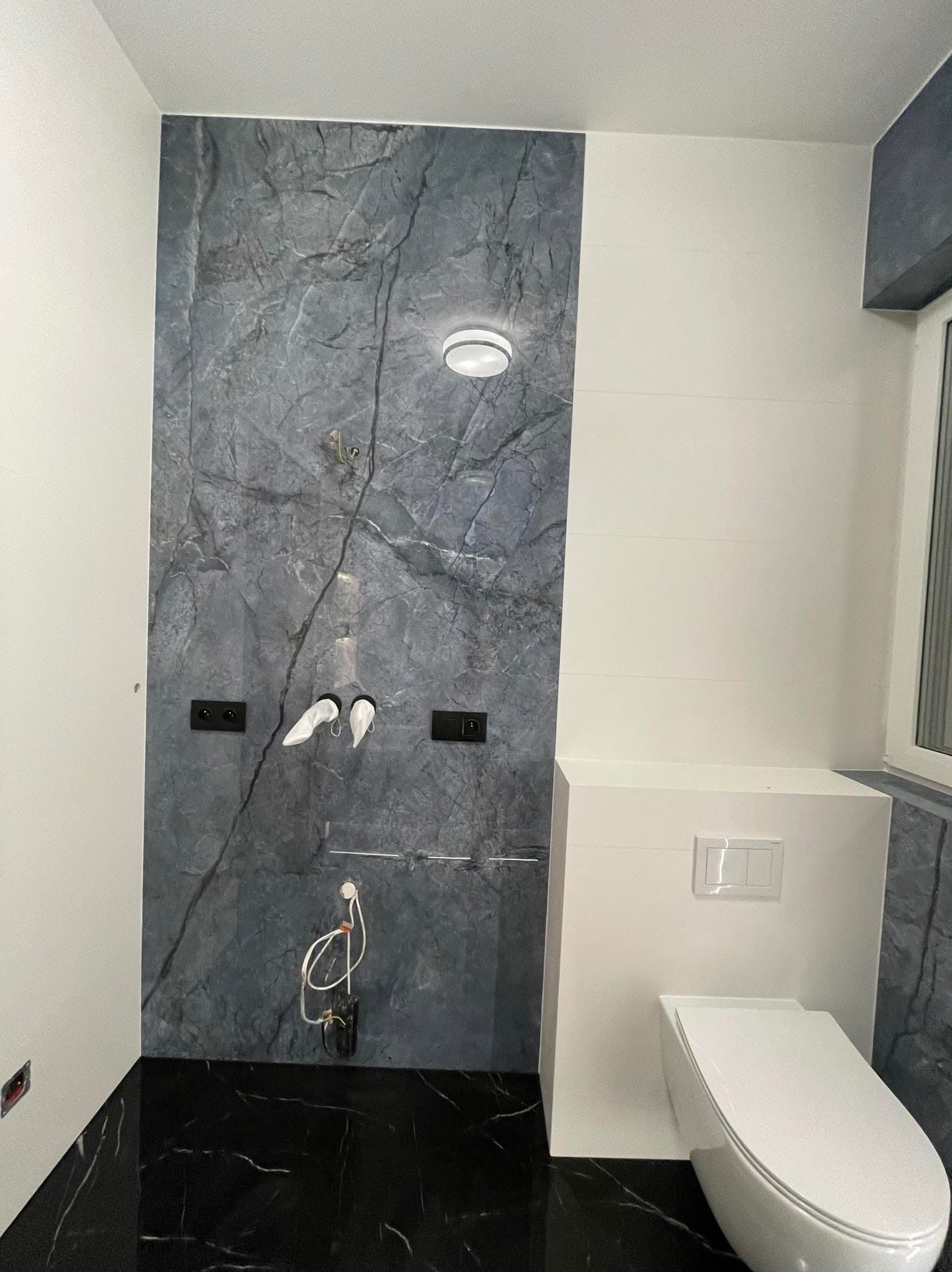 Nowoczesna łazienka w trakcie wykańczania, z marmurową płytą ścienną, czarną podłogą imitującą marmur, podwieszaną toaletą i przygotowanymi instalacjami wodno-kanalizacyjnymi.