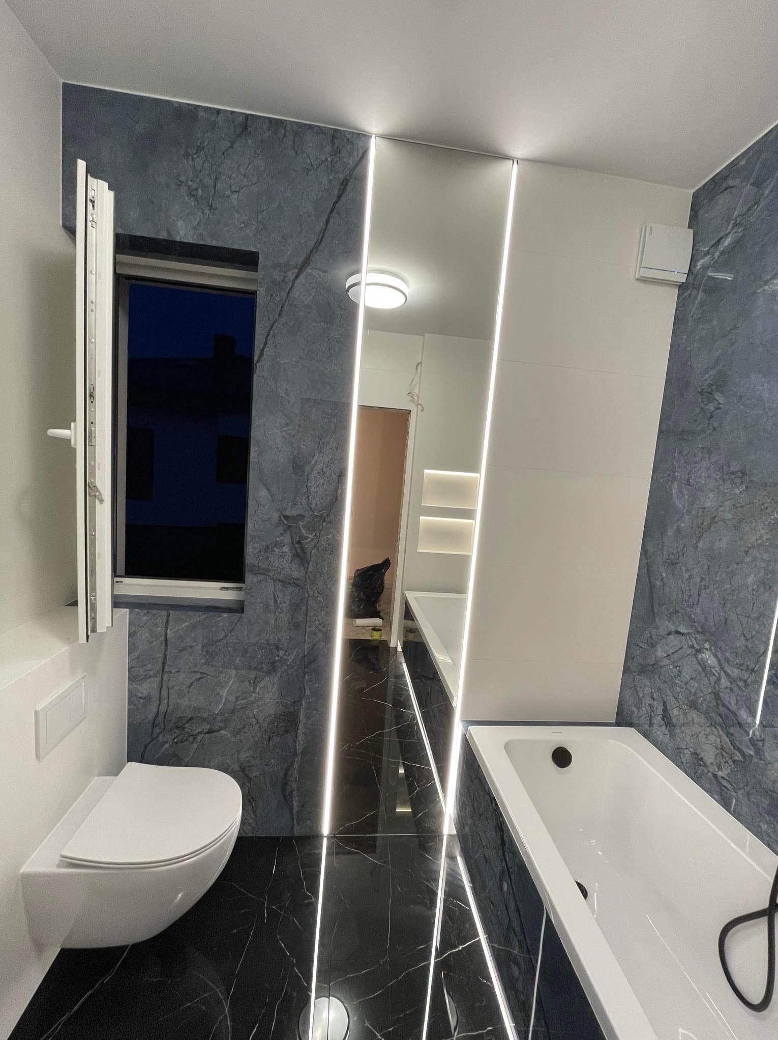 Nowoczesna łazienka z płytkami imitującymi marmur, podświetlanymi pionowymi pasami LED, wiszącą toaletą i wanną.