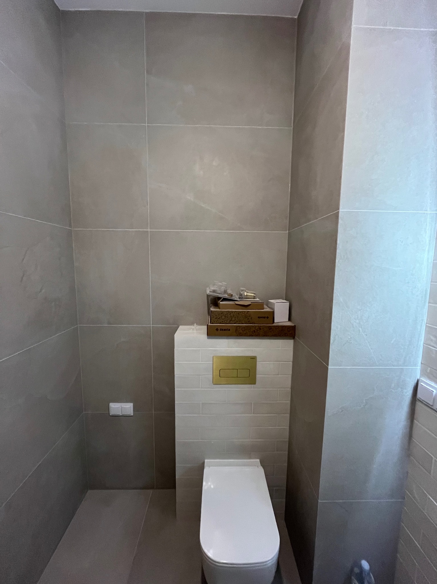 Nowoczesna, minimalistyczna toaleta w szaro-białej łazience z płytkami wielkoformatowymi i podtynkową spłuczką z złotym przyciskiem, na której leżą akcesoria montażowe w kartonie.