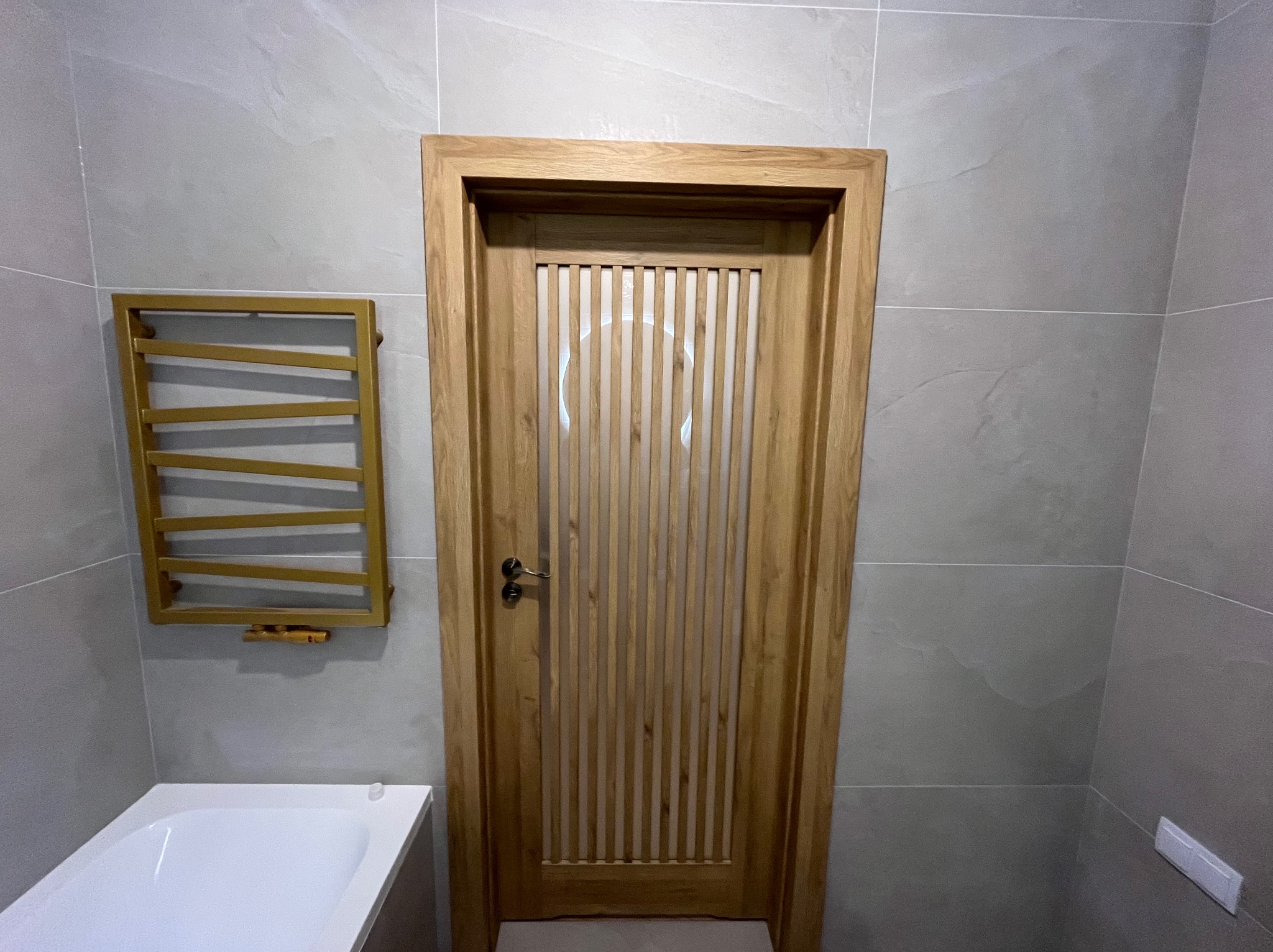 Wykończona łazienka z drewnianą, lamelową stolarką drzwiową, grzejnikiem drabinkowym w złotym kolorze i szarymi płytkami imitującymi kamień na ścianach.