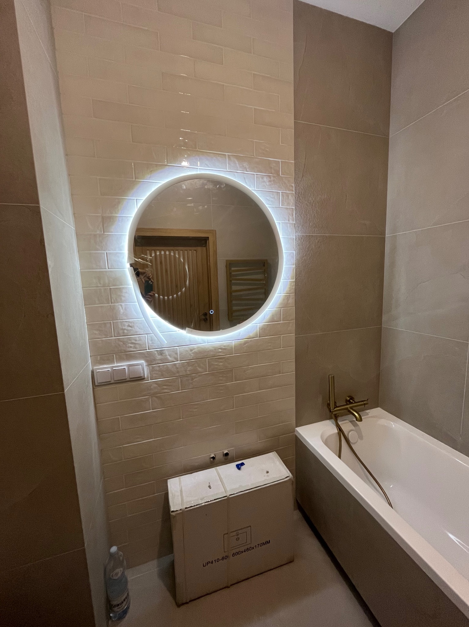 Nowoczesna łazienka w trakcie wykańczania z okrągłym lustrem LED, beżowymi płytkami imitującymi cegłę, złotą armaturą i kartonem po produkcie na podłodze.