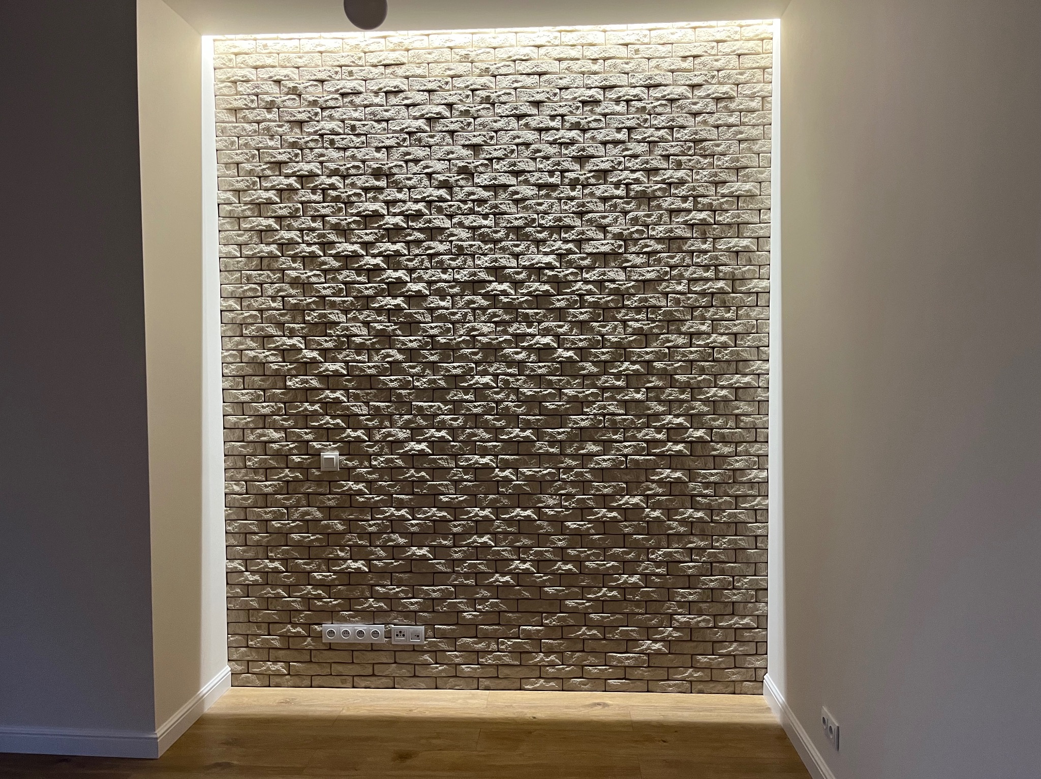 Ściana wykończona dekoracyjną cegłą z podświetleniem LED, widoczne gniazdka elektryczne i włącznik światła, drewniana podłoga