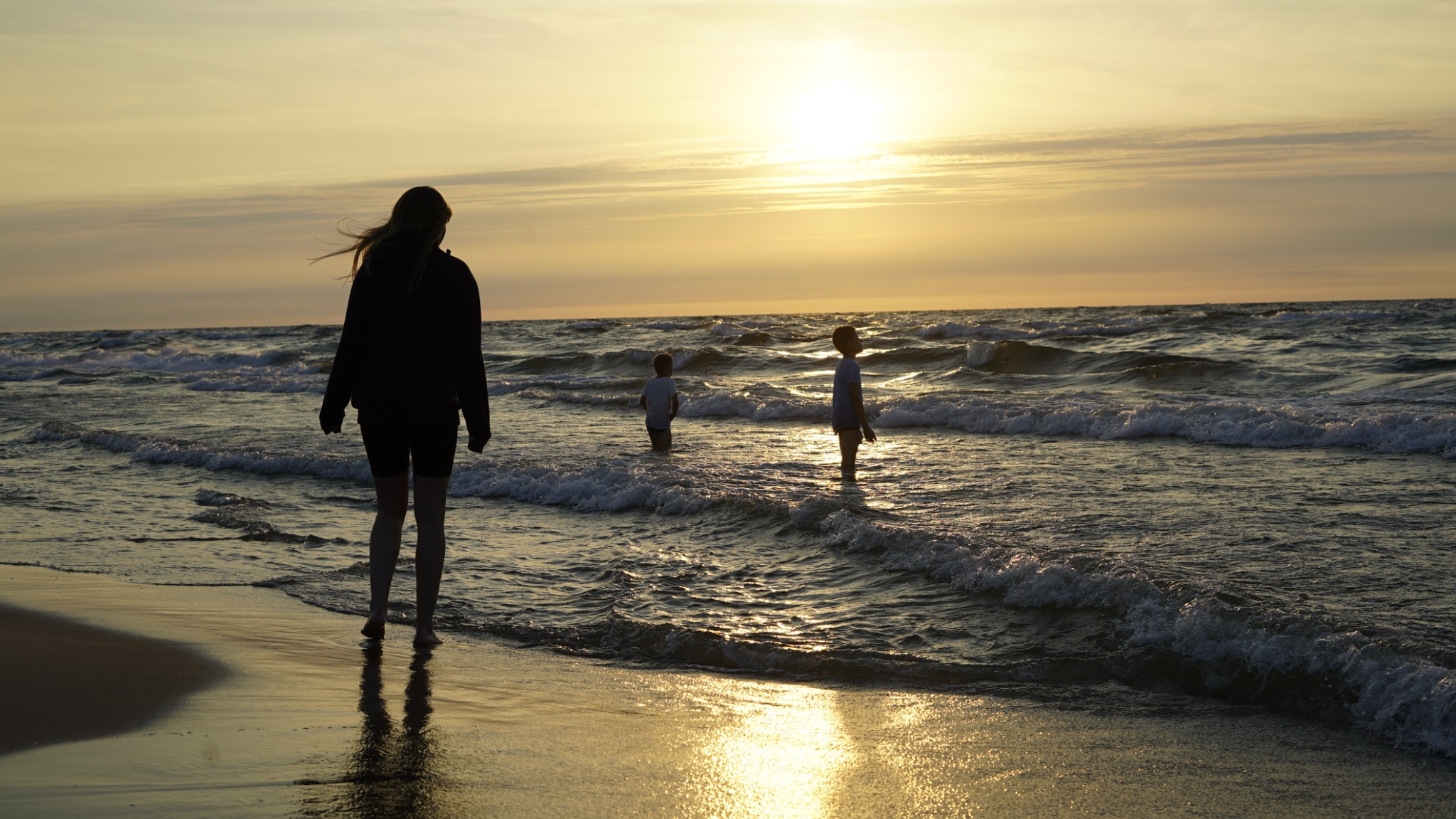 Sylwetka kobiety stojącej na brzegu morza o zachodzie słońca, w oddali dwoje dzieci brodzących w wodzie, fale rozbijają się o brzeg.