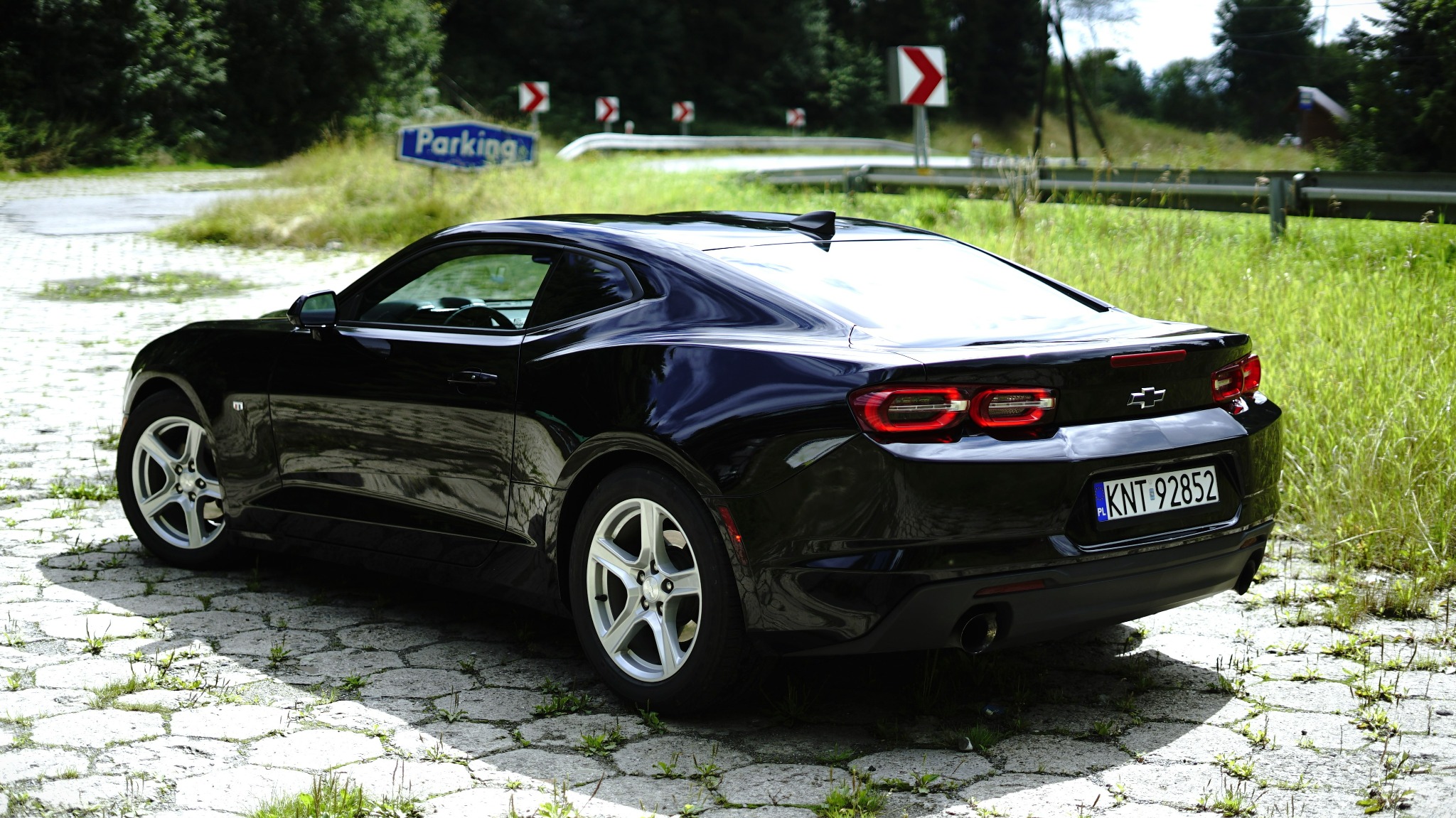 Czarny samochód Chevrolet Camaro zaparkowany na brukowanej drodze, z widocznymi znakami drogowymi i zielenią w tle.