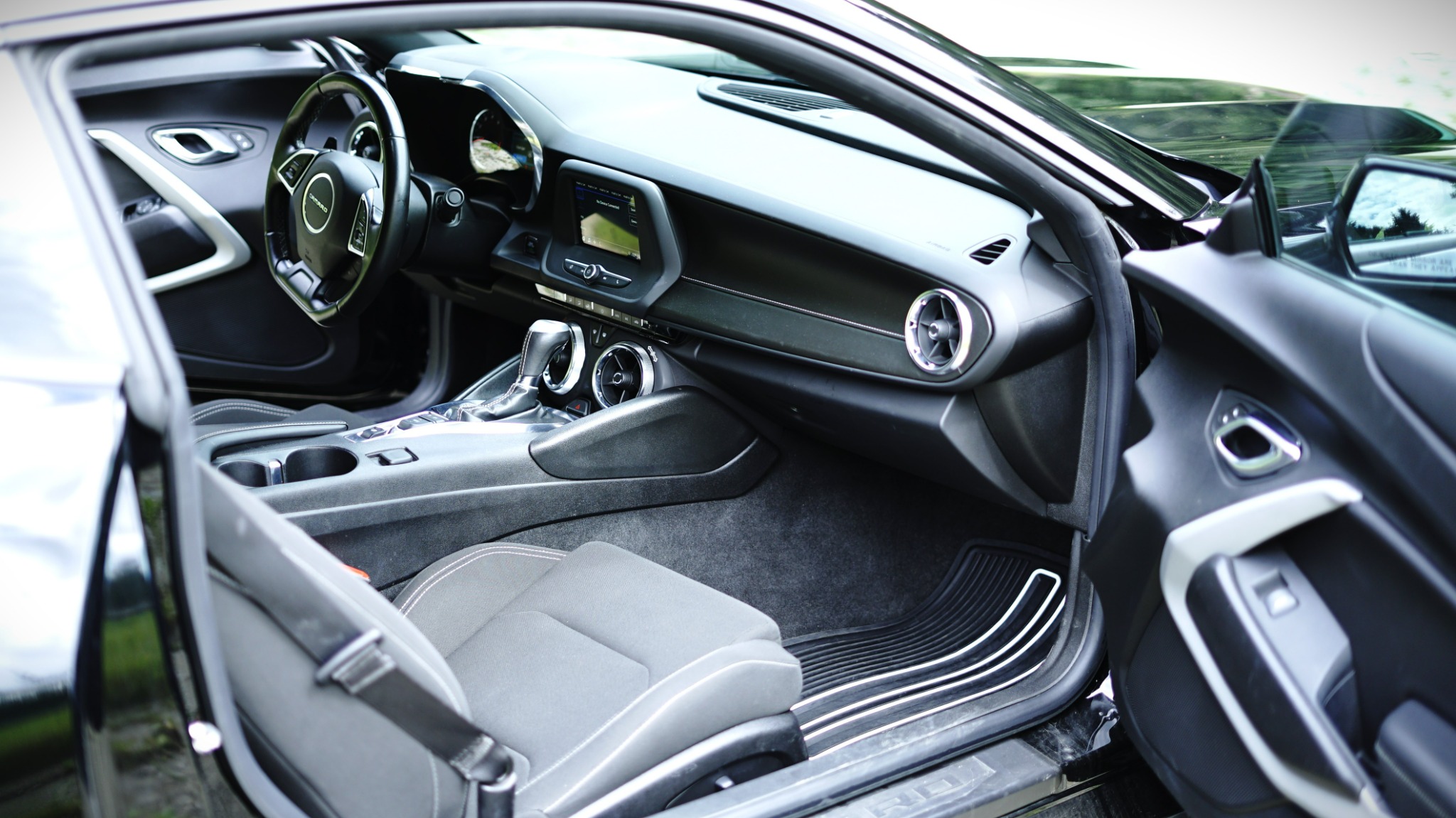 Wnętrze czarnego samochodu sportowego Camaro z otwartymi drzwiami, widoczny fotel kierowcy, deska rozdzielcza z ekranem dotykowym i srebrnymi akcentami oraz kierownica z logo Chevrolet.