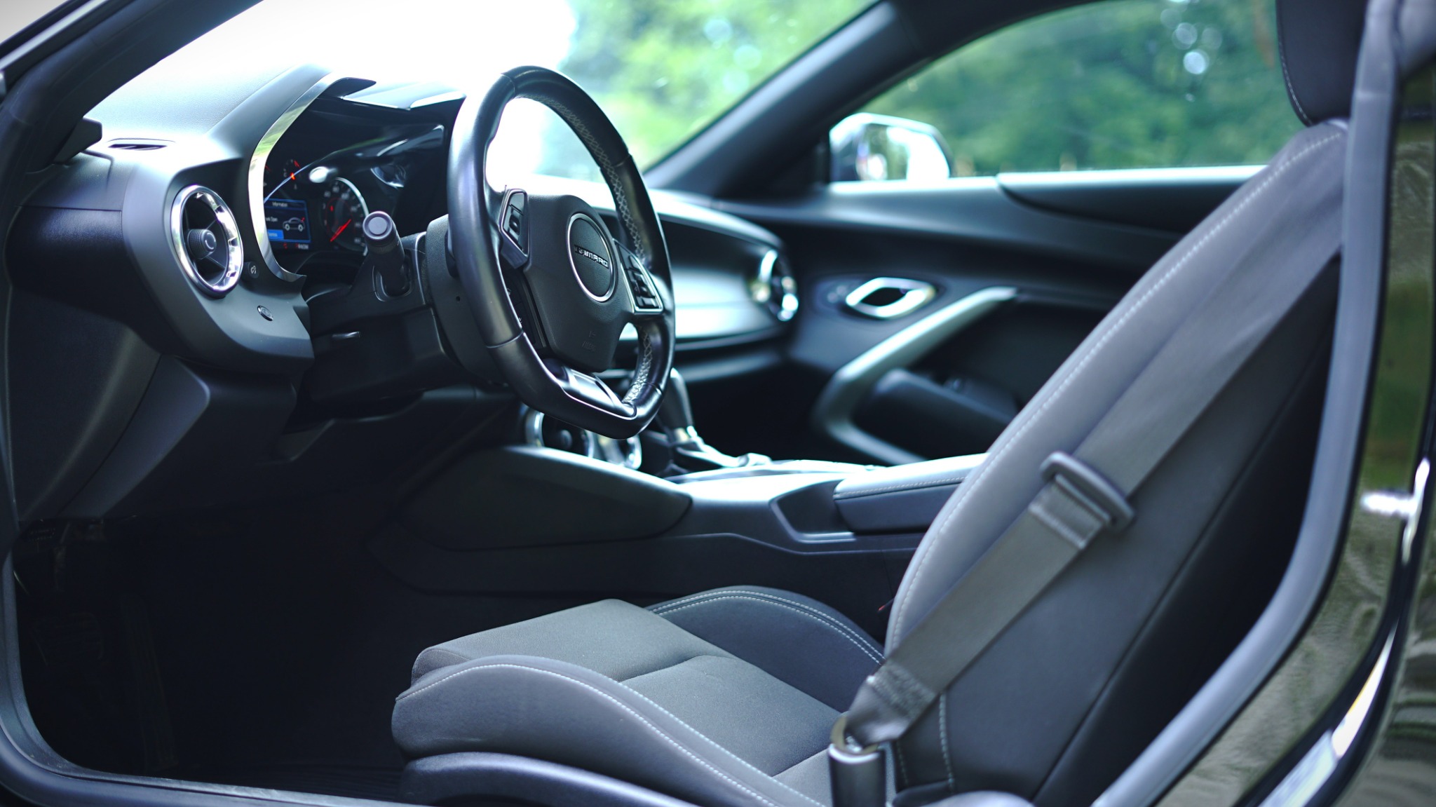 Wnętrze samochodu Chevrolet Camaro z otwartymi drzwiami, widoczny kokpit, kierownica z logo, szare fotele z białymi przeszyciami i pas bezpieczeństwa.