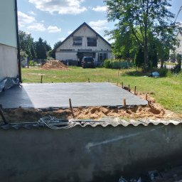 Adamczuk Paweł - Wylewka betonowa fundamentu domu jednorodzinnego w trakcie realizacji. Widoczny pracownik, szalunek i teren budowy z domem w tle.