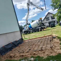Betoniarka z pompą podaje beton na przygotowane zbrojenie pod wylewkę fundamentu budynku z ocieploną elewacją. Widoczny pracownik i dom w tle.
