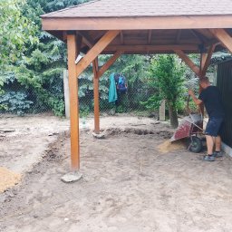 Adamczuk Paweł - Budowa drewnianej altany: mężczyzna rozprowadza piasek wewnątrz konstrukcji. Widoczne narzędzia, ogrodzenie i zieleń w tle. Prace ziemne w toku.