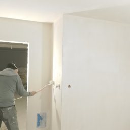 Adamczuk Paweł - Malowanie ścian wałkiem w jasnym wnętrzu. Widoczny fragment sylwetki malarza i przygotowane do malowania ściany z widocznymi śladami po taśmie malarskiej.