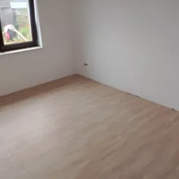 Jasne panele podłogowe ułożone w pokoju z białymi ścianami i oknem. Widoczne gniazdka elektryczne na ścianie. Minimalistyczne wykończenie wnętrza.