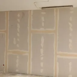 Ściana z płyt gipsowo-kartonowych, z widocznymi łączeniami i miejscami po szpachlowaniu. W tle widoczna lampa sufitowa i fragment konstrukcji dachu.