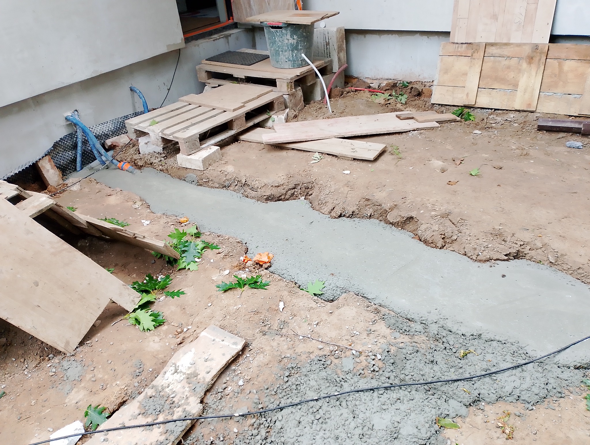 Wylany fundament pod budynek. Widoczne palety, rury i wykopane fundamenty. Na ziemi leżą liście i kawałki drewna. Fundament świeżo wylany, jeszcze mokry.