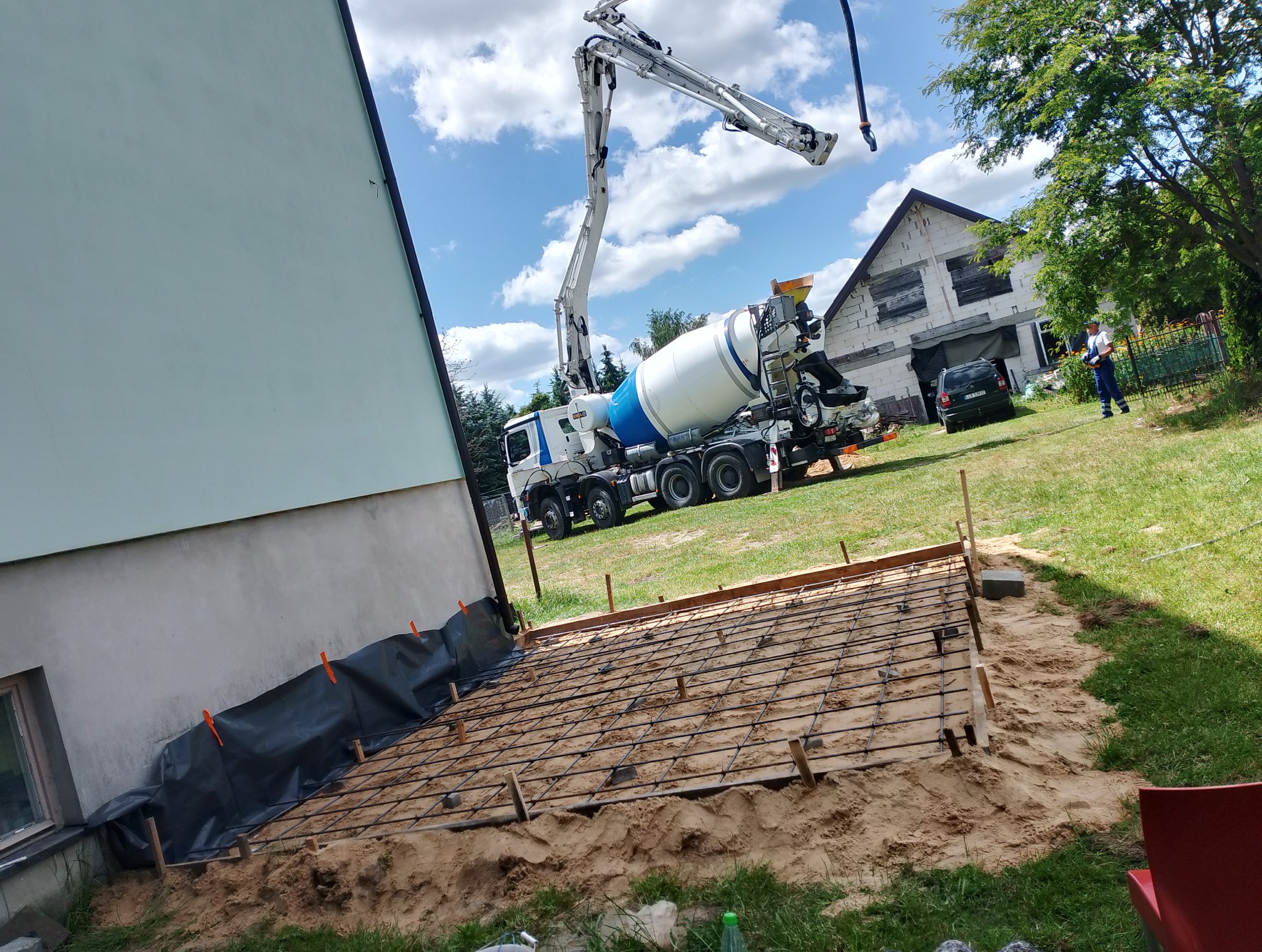 Betoniarka z pompą podaje beton na przygotowane zbrojenie pod wylewkę fundamentu budynku z ocieploną elewacją. Widoczny pracownik i dom w tle.