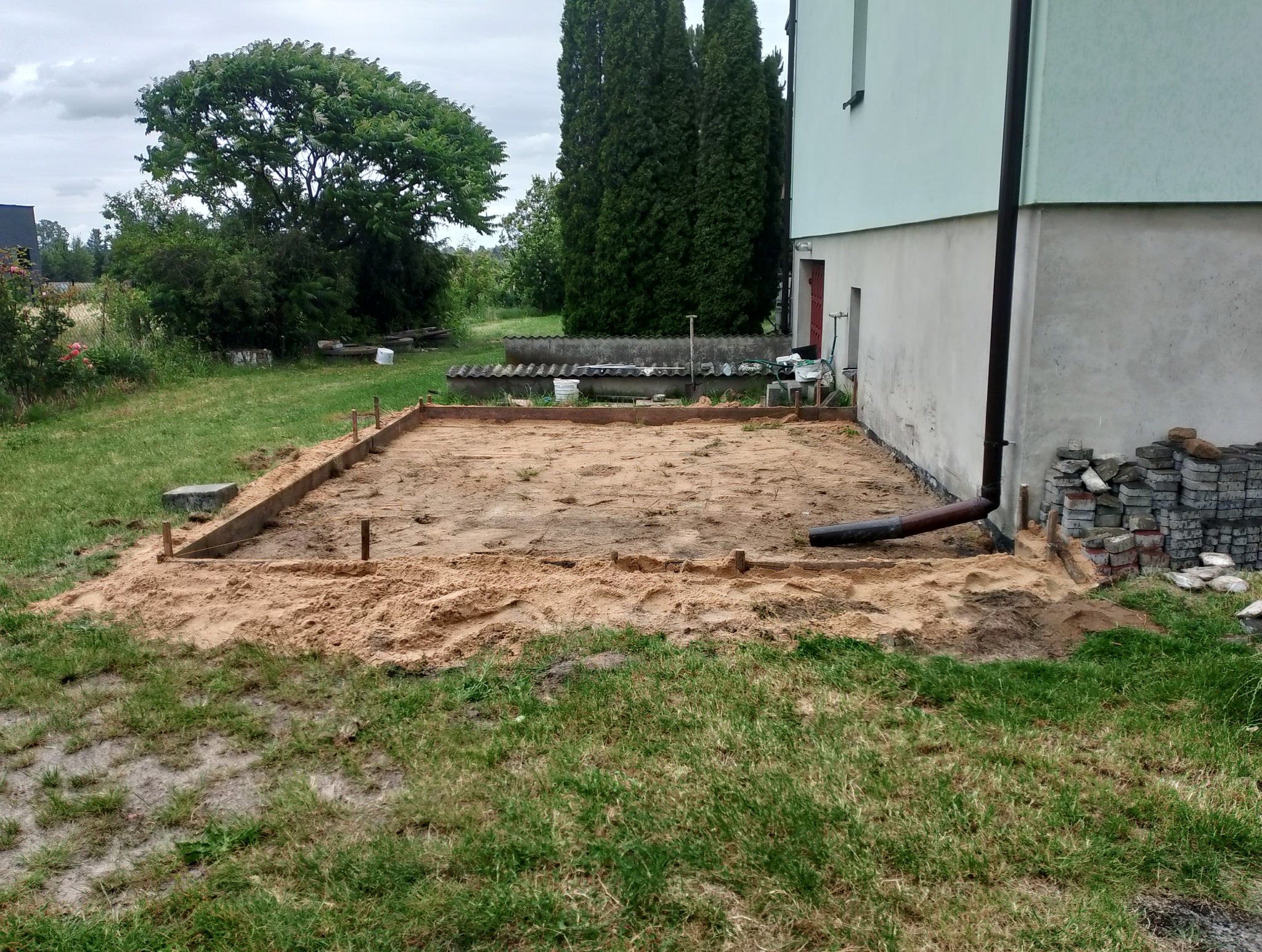 Przygotowanie terenu pod budowę tarasu: drewniany szalunek wypełniony piaskiem, widoczny fragment elewacji budynku i zieleń ogrodu w tle.