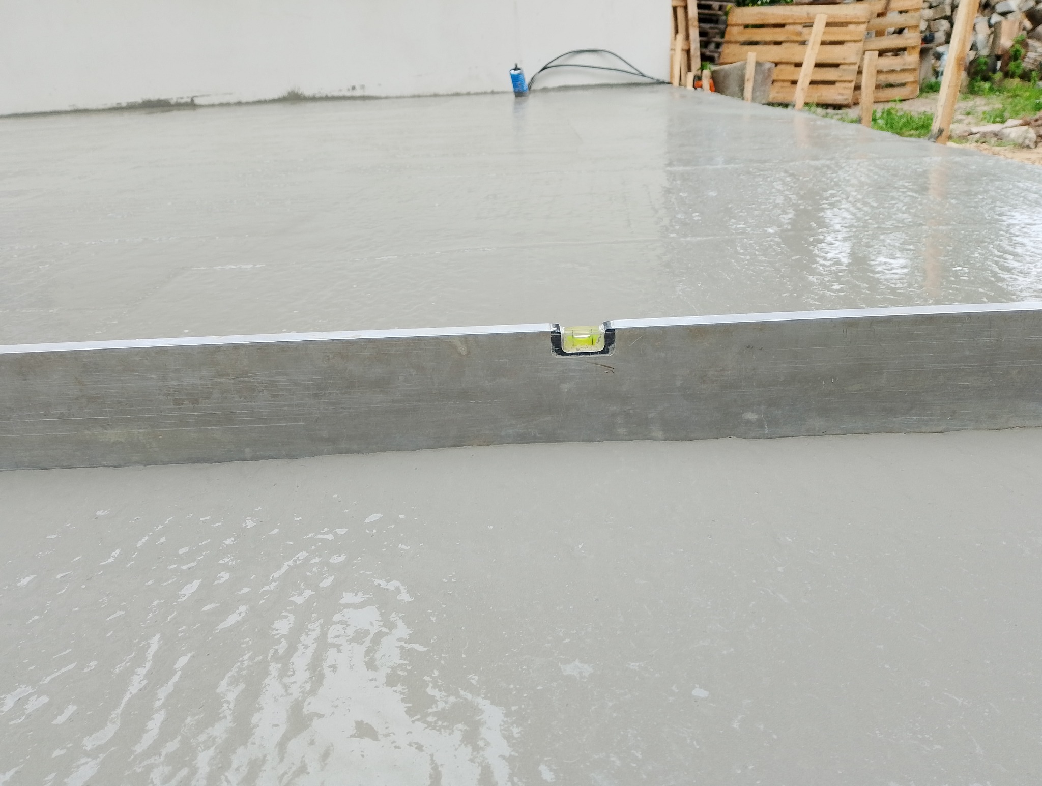 Wyrównywanie wylewki poziomnicą na budowie. Widoczne drewniane elementy szalunku i fragment ściany. Jasne, naturalne światło.