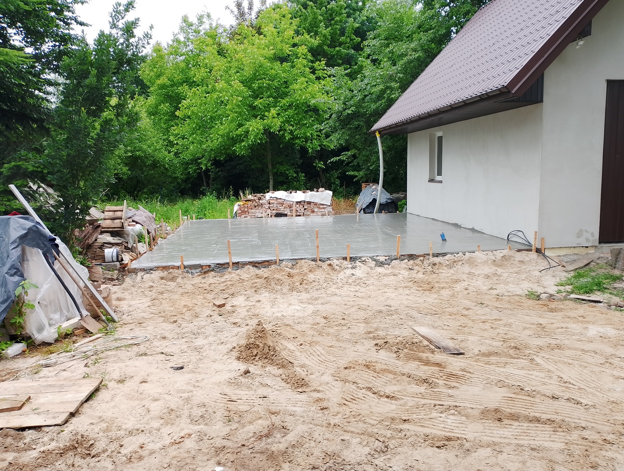 Wylany fundament pod budynek z widoczną konstrukcją szalunkową i stertą cegieł w tle, otoczony piaszczystym terenem.