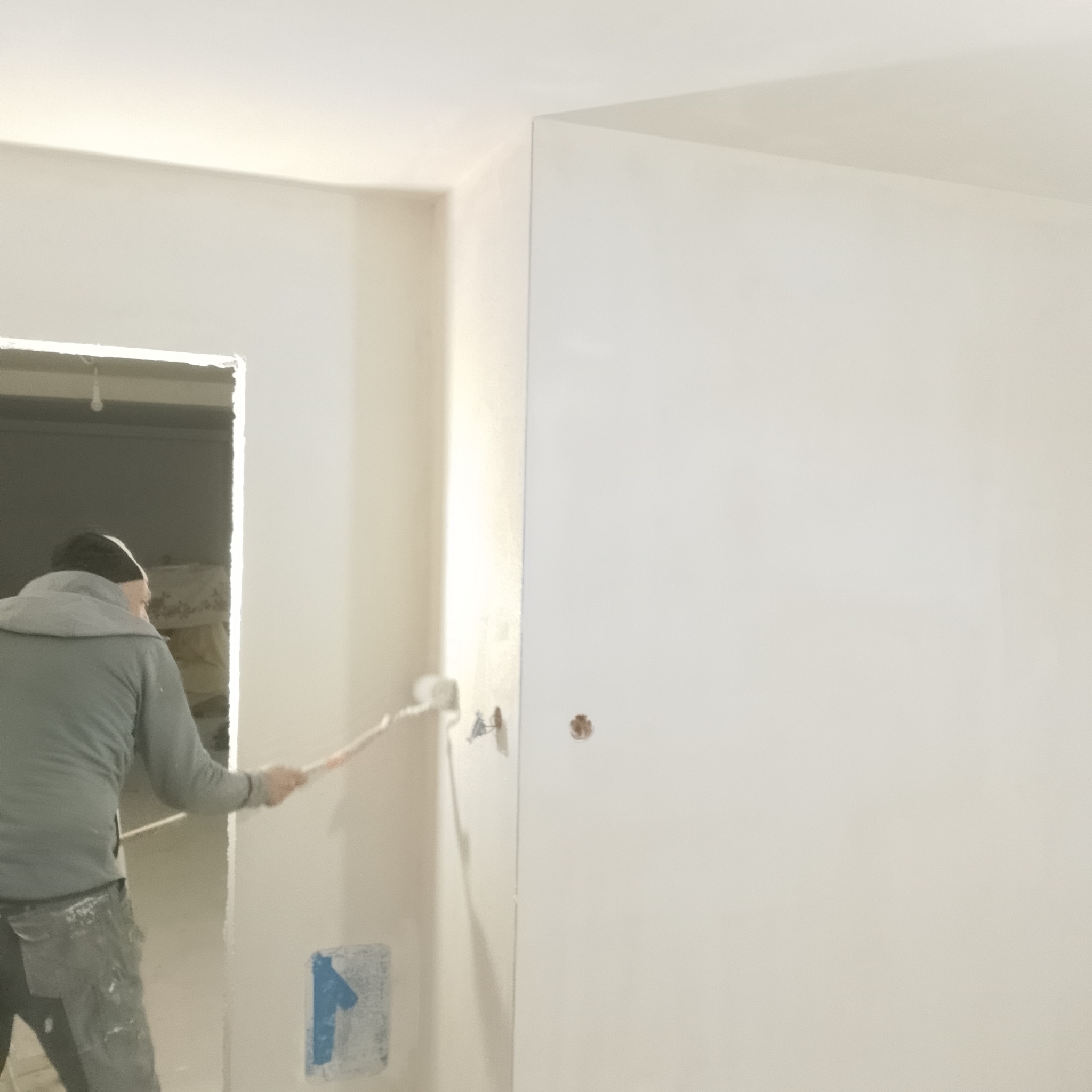 Malowanie ścian wałkiem w jasnym wnętrzu. Widoczny fragment sylwetki malarza i przygotowane do malowania ściany z widocznymi śladami po taśmie malarskiej.