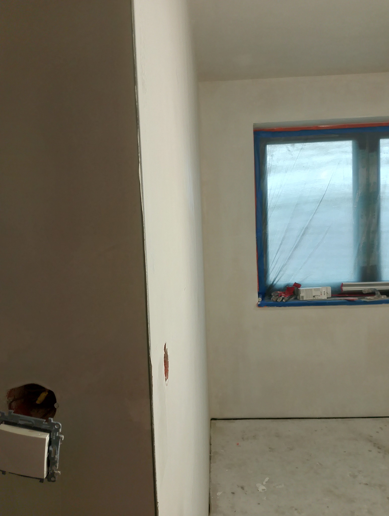 Wnętrze w trakcie gładzenia ścian, z widocznym oknem zabezpieczonym folią i puszką elektryczną. Ściany przygotowane pod malowanie, surowy beton na podłodze.