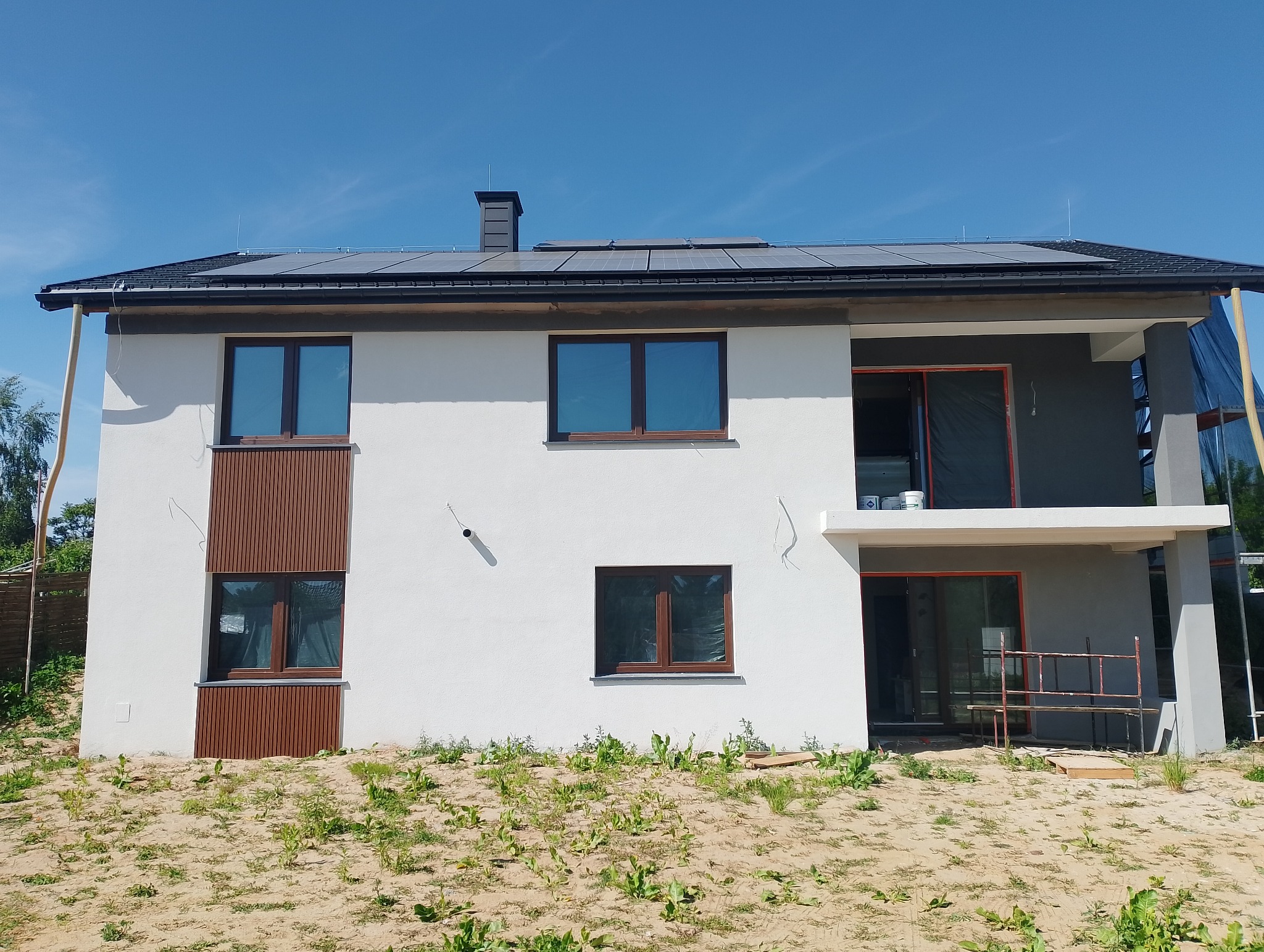 Dwukondygnacyjny dom z panelami słonecznymi na dachu, częściowo wykończoną elewacją (biel i szarość), brązowymi ramami okiennymi i dekoracyjnymi panelami.