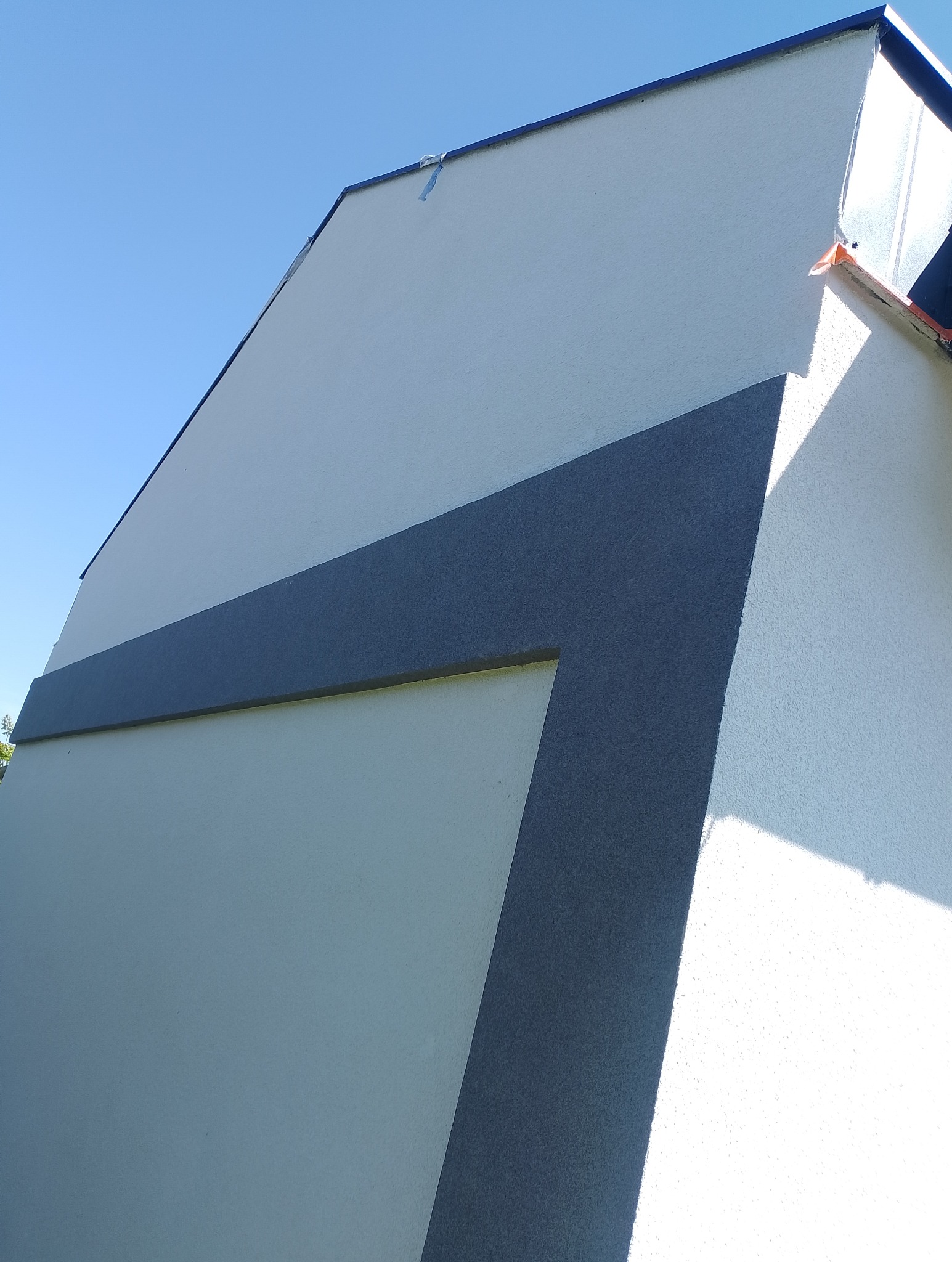 Elewacja budynku z dekoracyjnym, ciemnoszarym pasem, widok z dołu. Jasne tło nieba kontrastuje z bielą ścian. Nowoczesne wykończenie.