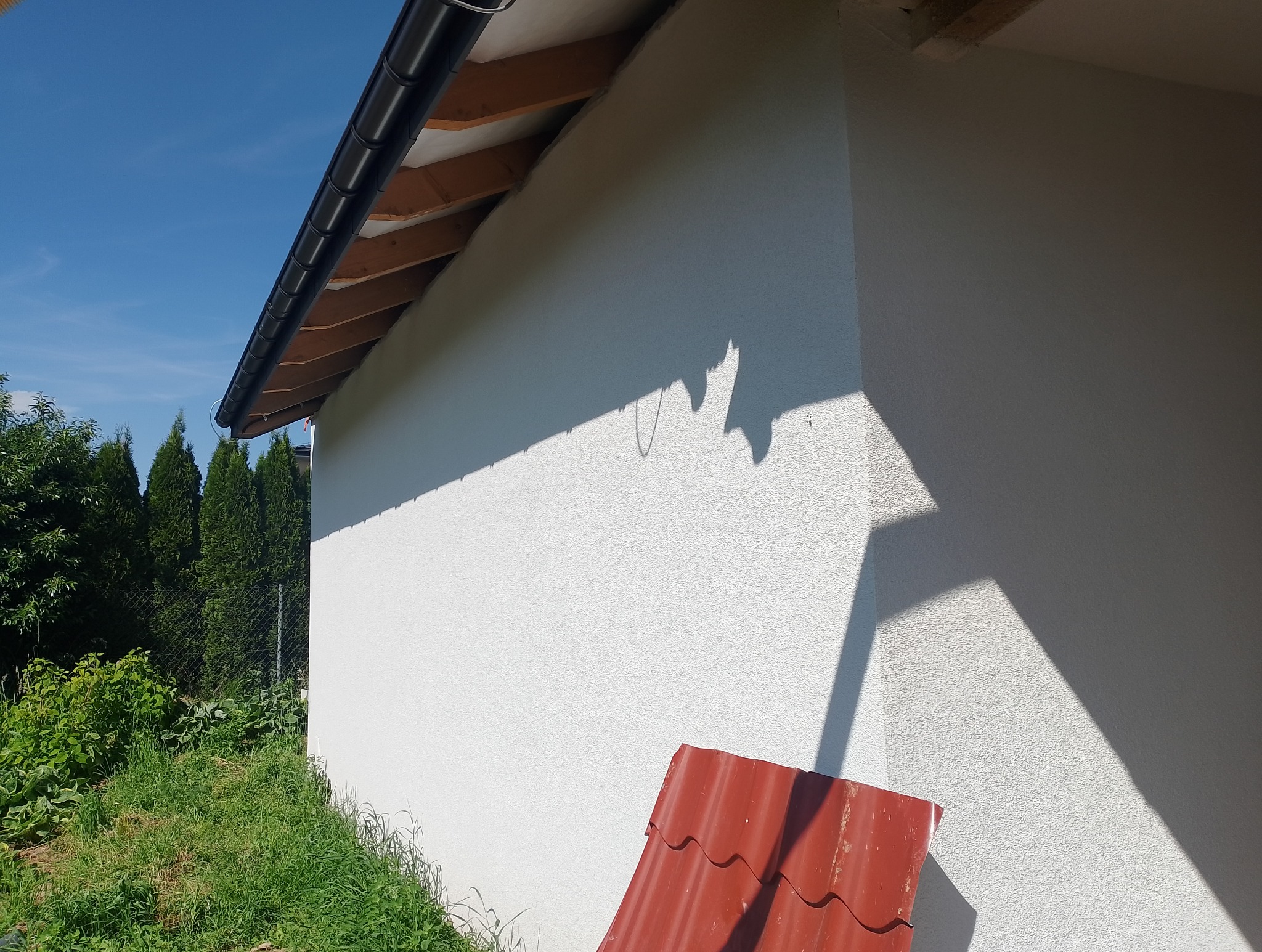 Biała elewacja budynku w słoneczny dzień, widoczny fragment dachu z rynną i zielenią. W rogu leży czerwona dachówka. Teksturowana powierzchnia elewacji.