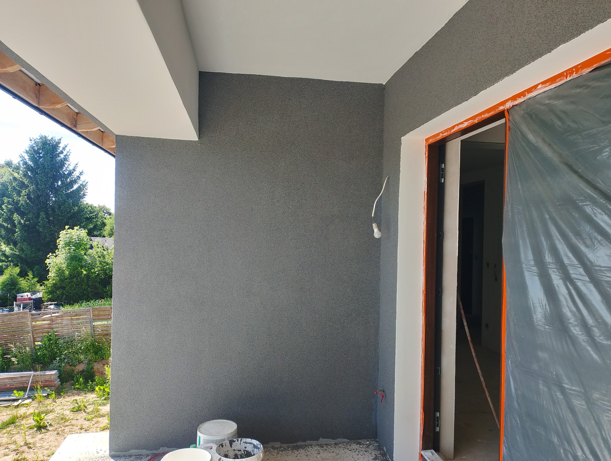 Szara elewacja zewnętrzna z efektem baranka, białe wykończenia okien i sufitu, zabezpieczone drzwi folią malarską, widok na zieleń z zewnątrz.