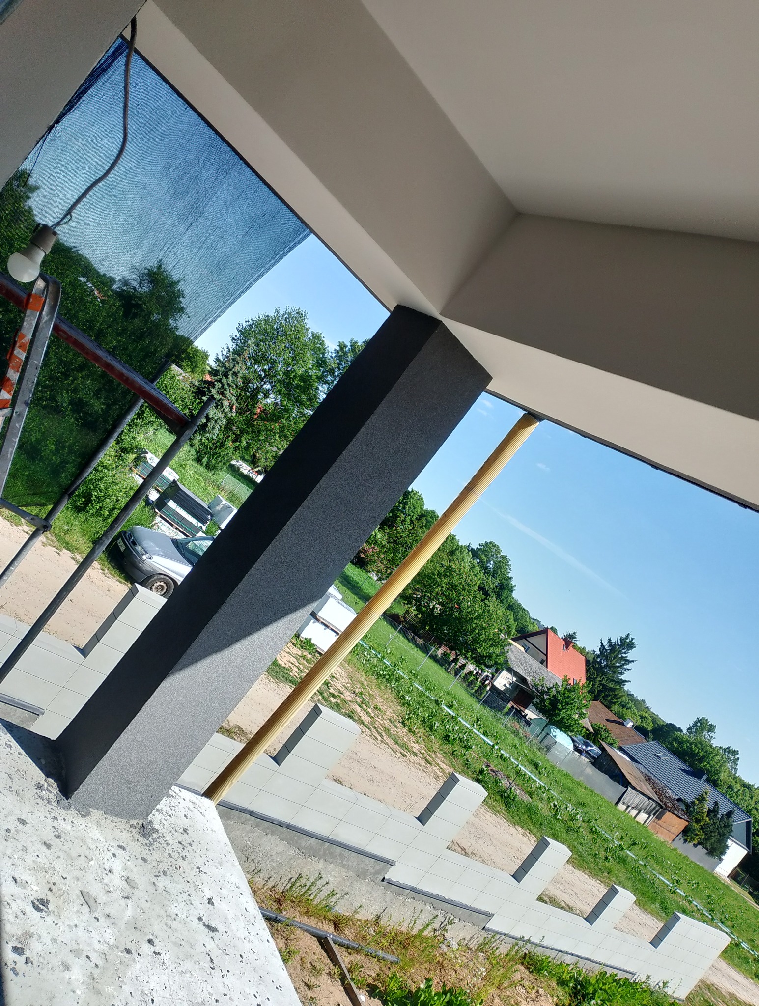 Świeżo pomalowany, grafitowy filar balkonu w surowym stanie, z widoczną elewacją budynku w tle i elementami rusztowania. Widok z dołu pod kątem.