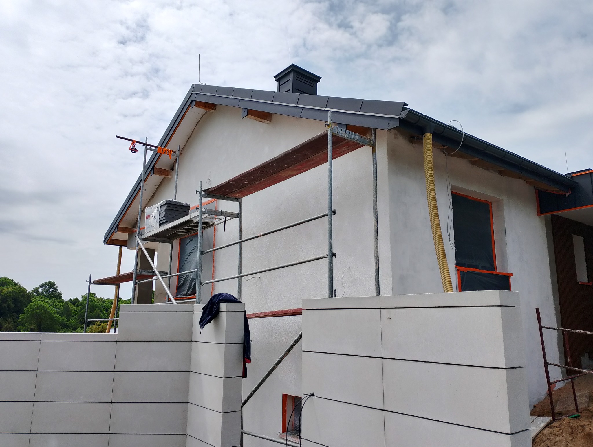 Dom jednorodzinny w trakcie ocieplania, widoczne rusztowania i zabezpieczone okna. Elewacja w trakcie prac wykończeniowych, widoczny fragment ocieplonej ściany.