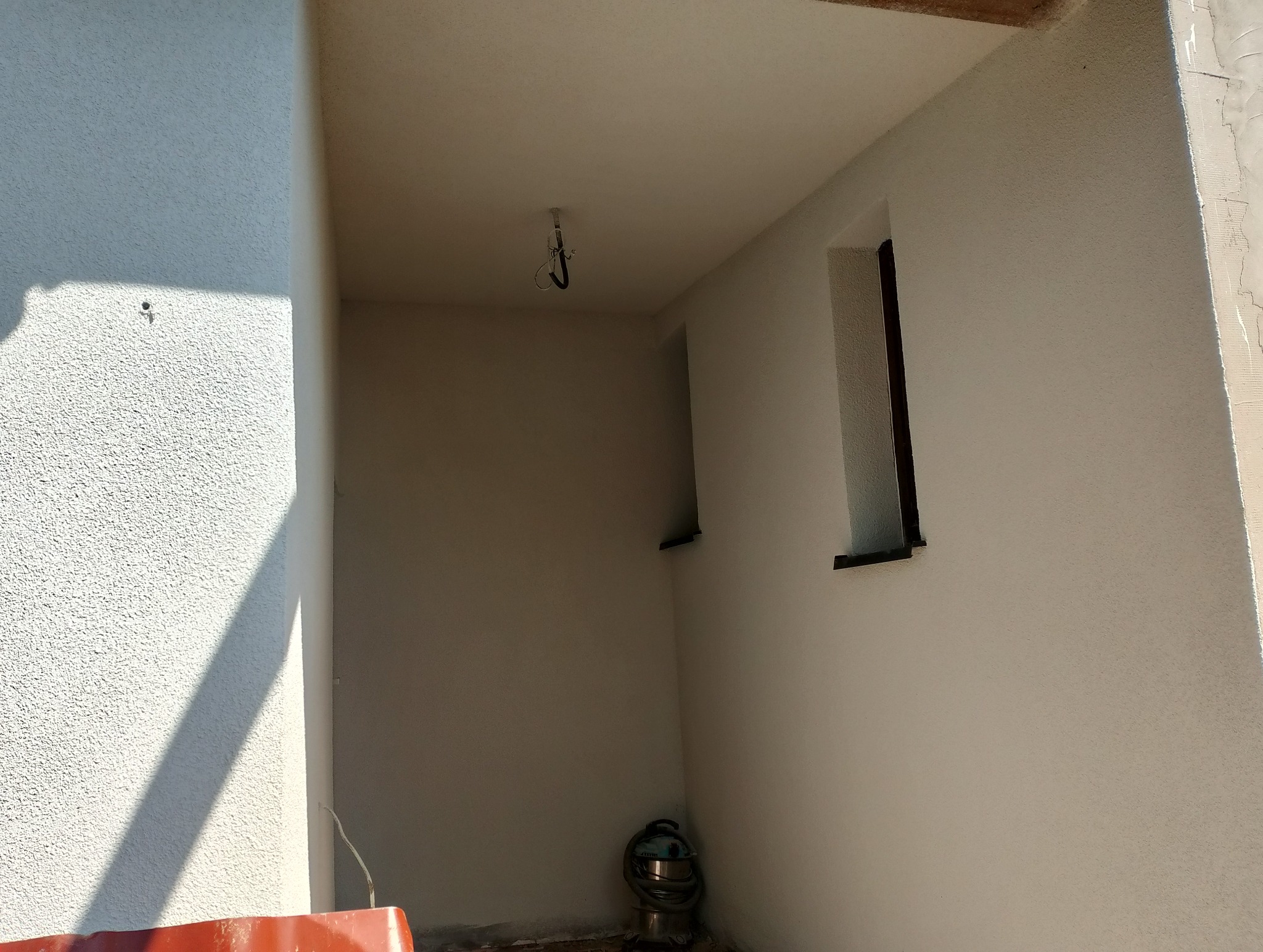 Wnętrze w trakcie wykończenia: ściany z fakturą, okna z ciemnymi ramami, widoczny odkurzacz przemysłowy. Oświetlenie dzienne podkreśla strukturę tynku.