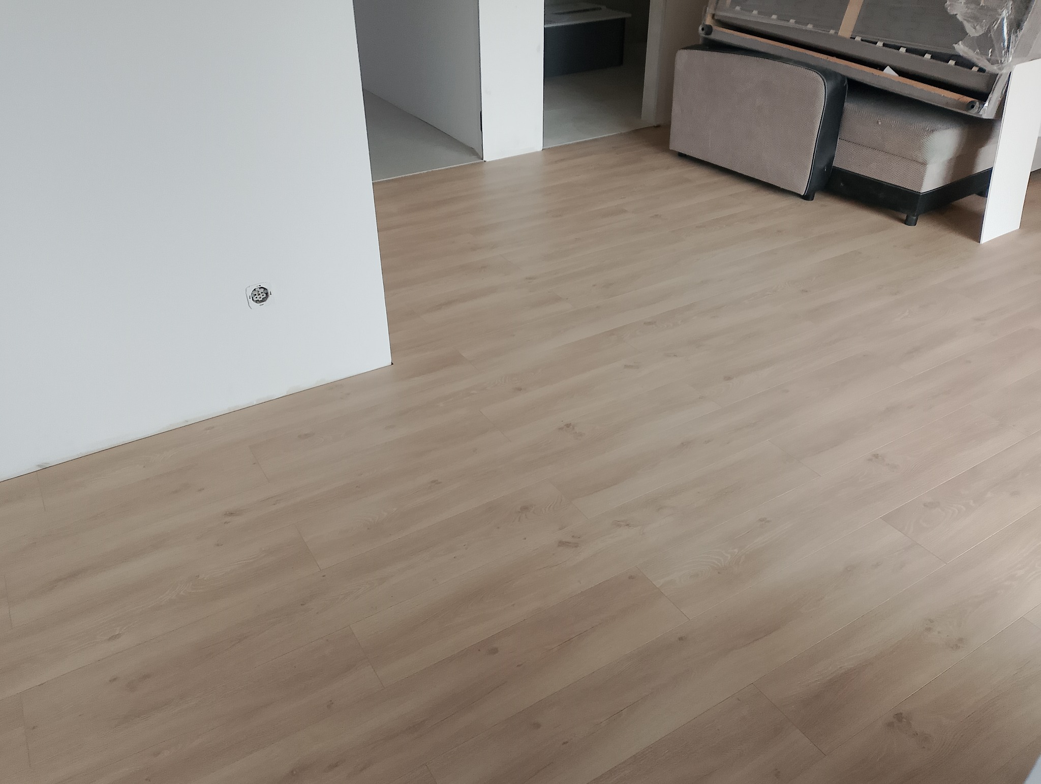 Jasne panele podłogowe w nowym wnętrzu, widoczna ściana z gniazdkiem elektrycznym i fragment mebla w tle. Minimalistyczny design, proste linie, naturalne światło.