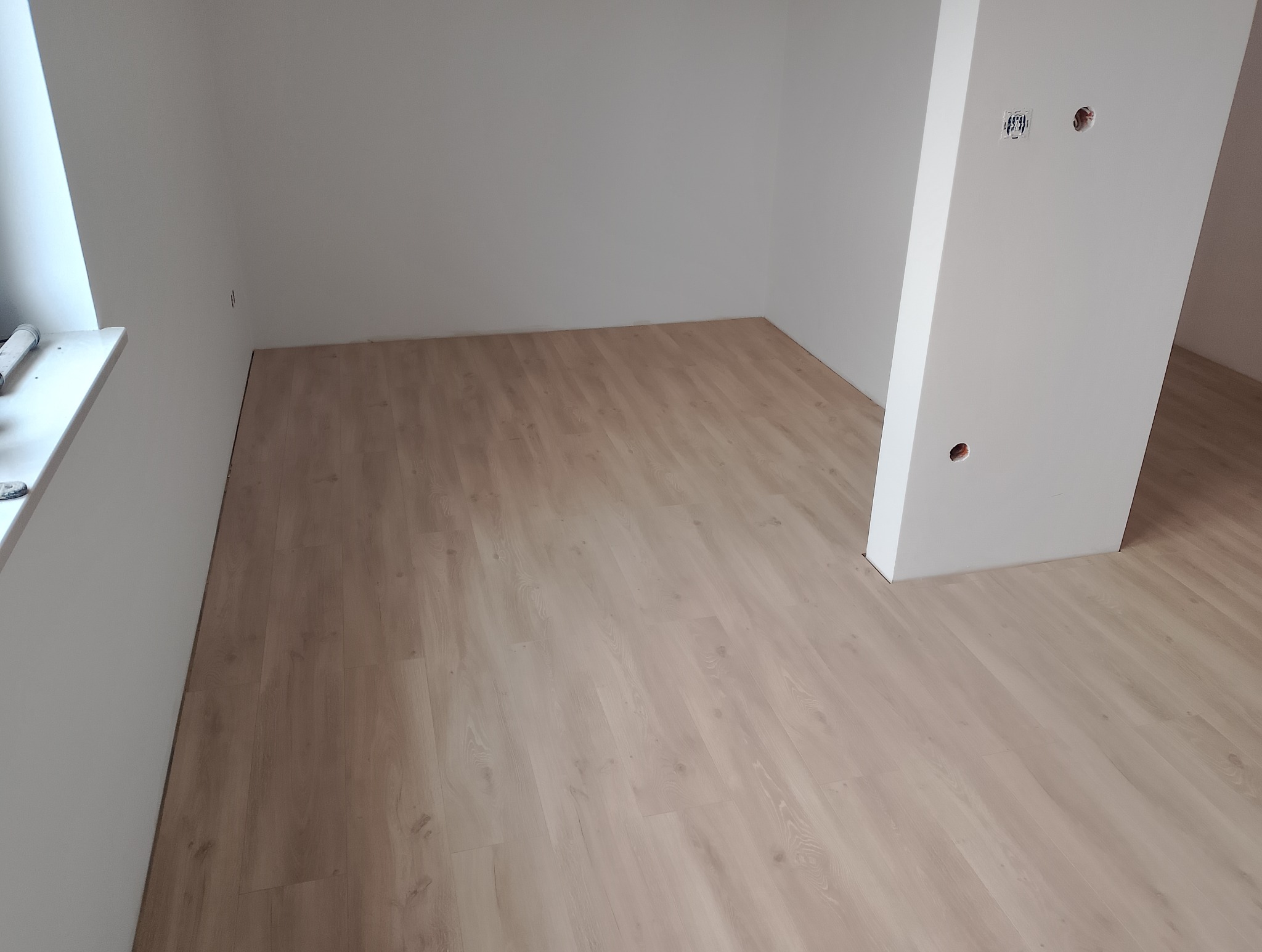 Jasne panele podłogowe ułożone w pustym, odświeżonym pokoju z białymi ścianami i widocznymi gniazdkami elektrycznymi. Minimalistyczne wykończenie wnętrza.