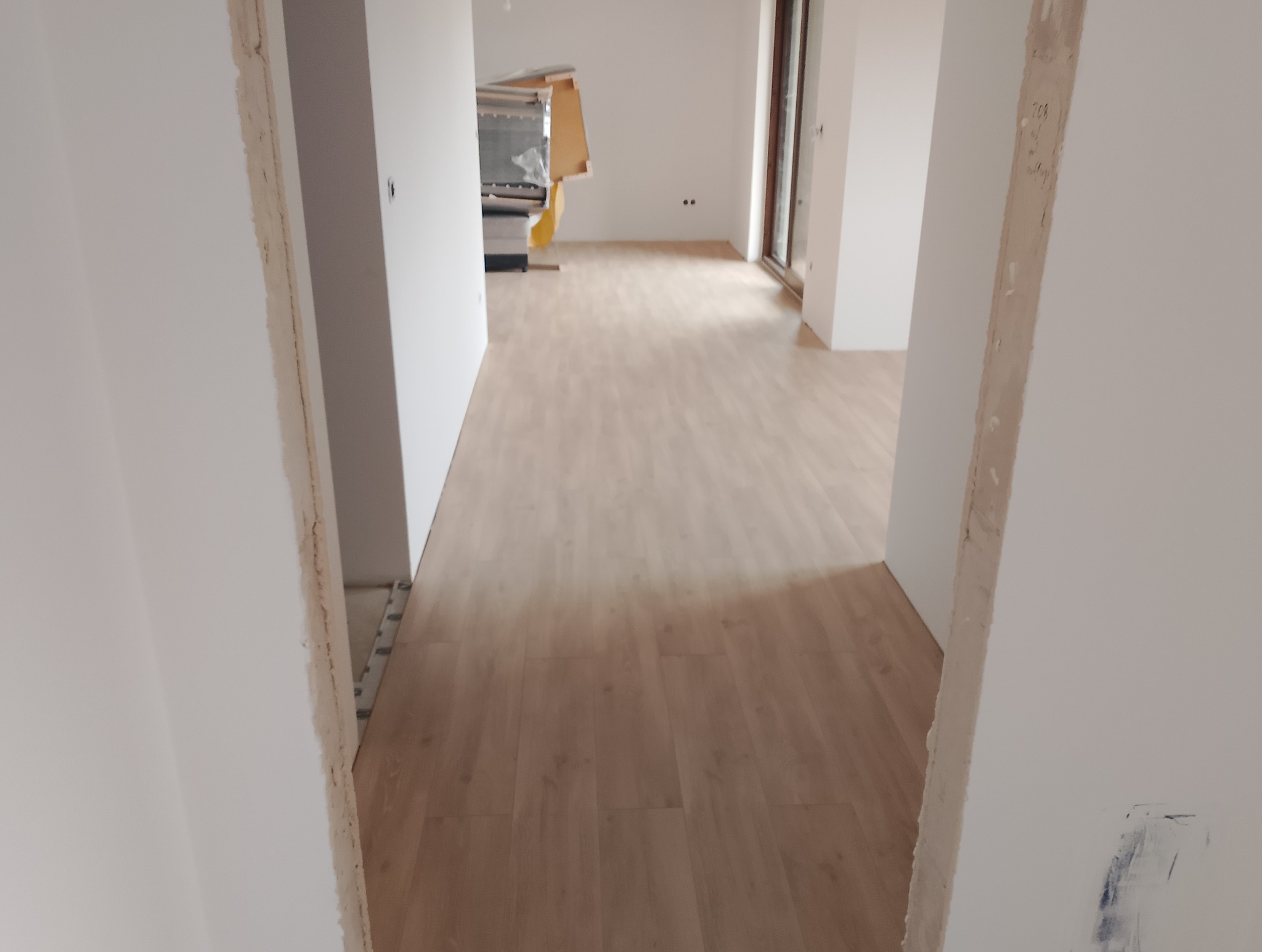 Jasne panele podłogowe ułożone w pustym, świeżo wyremontowanym pokoju. Widok przez surową futrynę drzwiową, ściany pomalowane na biało.