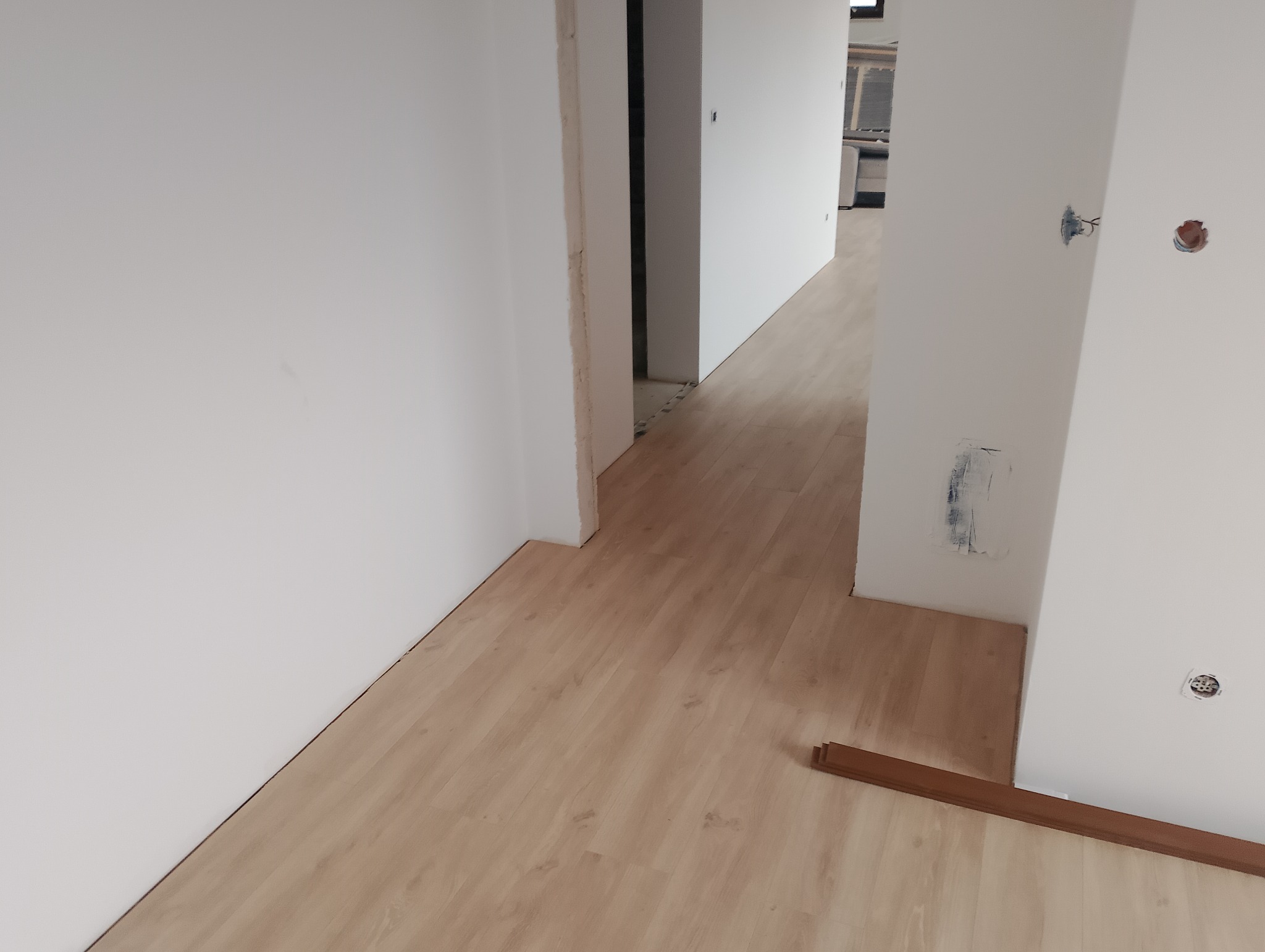 Jasne panele podłogowe ułożone w korytarzu z białymi ścianami. Widoczne niedokończone fragmenty przy ścianie i listwa przypodłogowa. Gniazdka elektryczne bez obudowy.