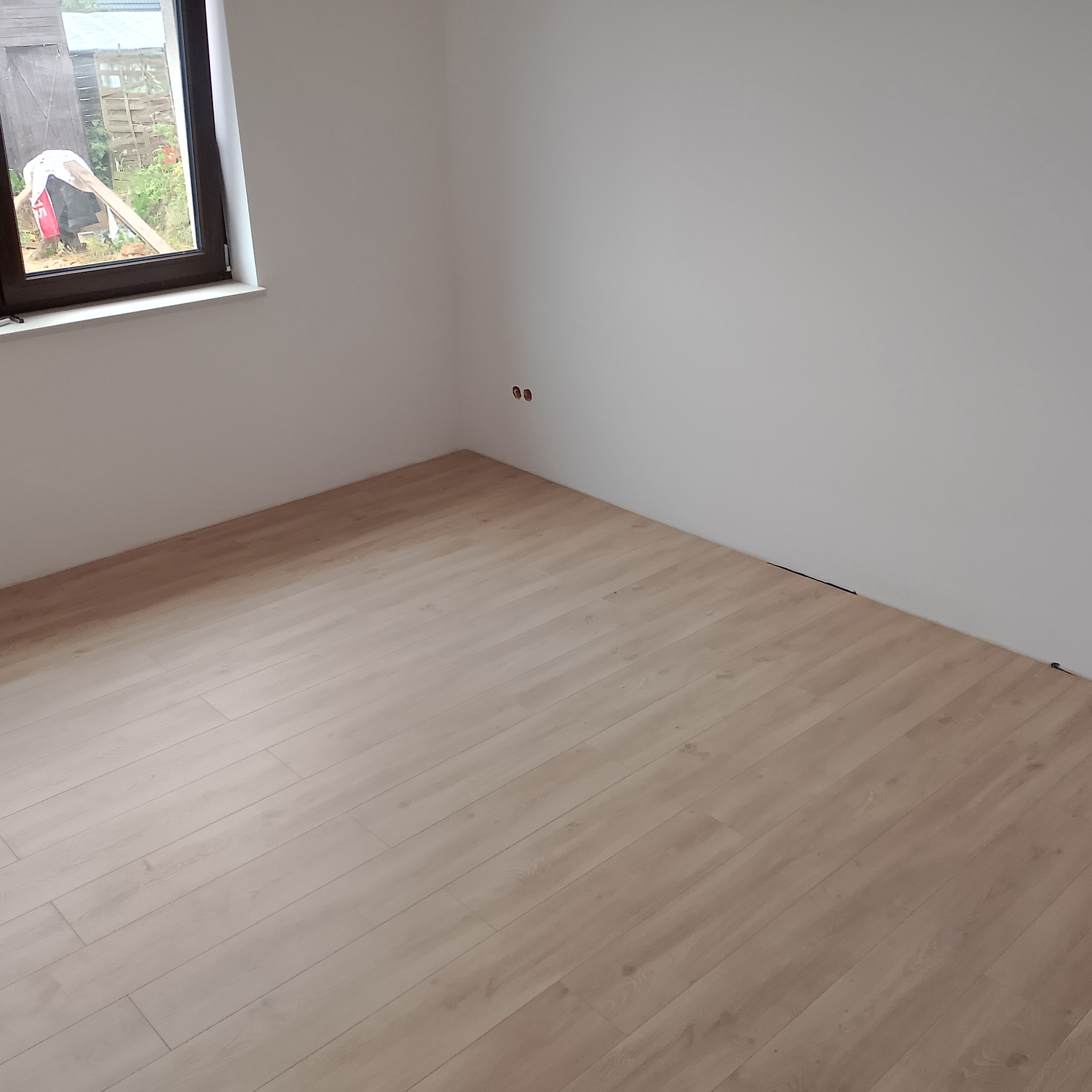 Jasne panele podłogowe ułożone w pokoju z białymi ścianami i oknem. Widoczne gniazdka elektryczne na ścianie. Minimalistyczne wykończenie wnętrza.