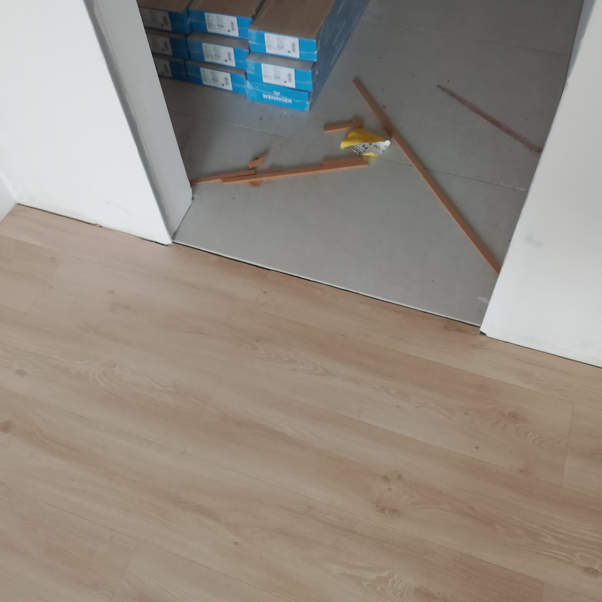 Jasne panele podłogowe ułożone w przejściu między pomieszczeniami. W tle widoczne kartony paneli i fragmenty listew. Wnętrze w trakcie prac wykończeniowych.