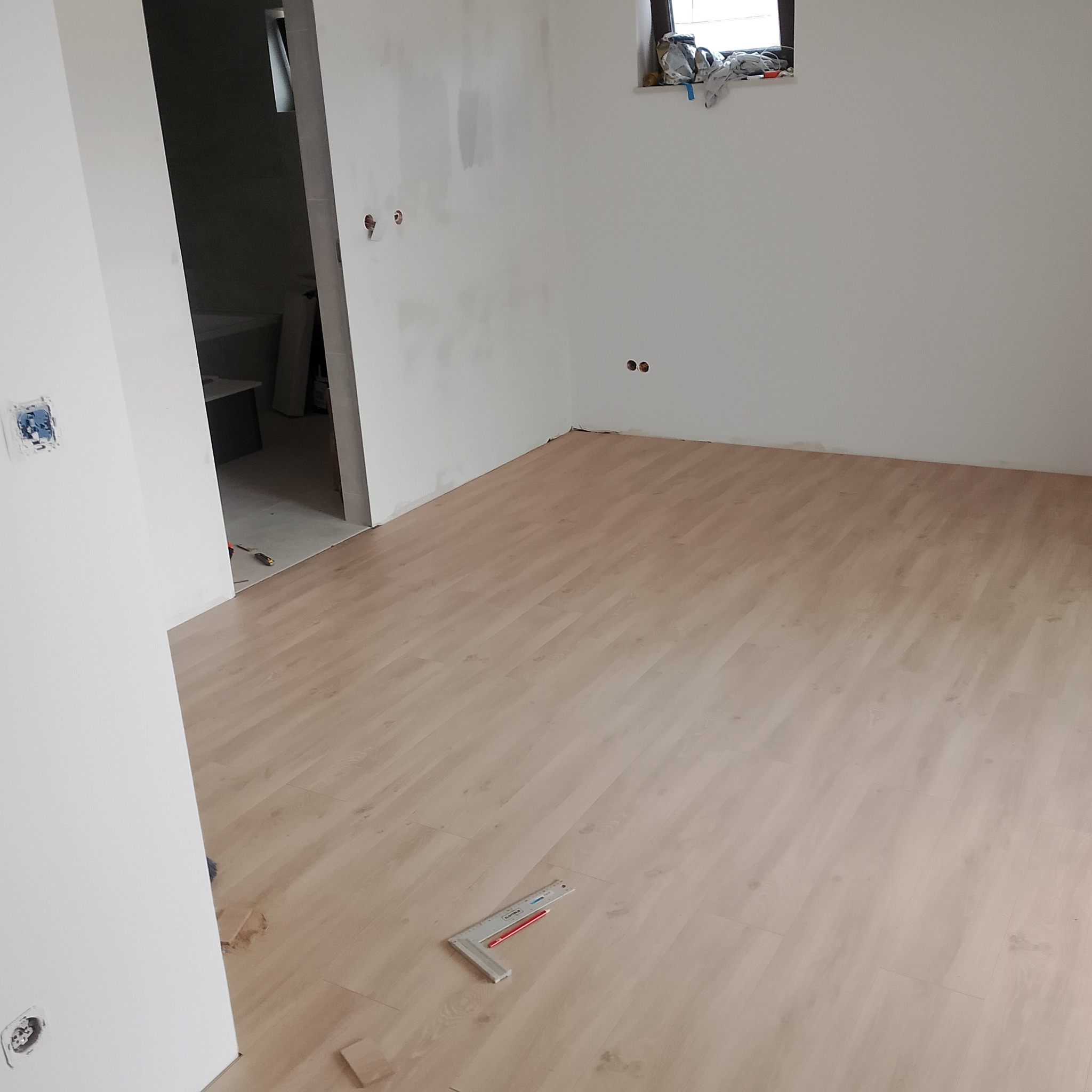 Jasne panele ułożone w pokoju, widoczne narzędzie (kątownik) i ołówek na podłodze. Ściany białe, w tle widoczne wejście do łazienki w trakcie remontu.