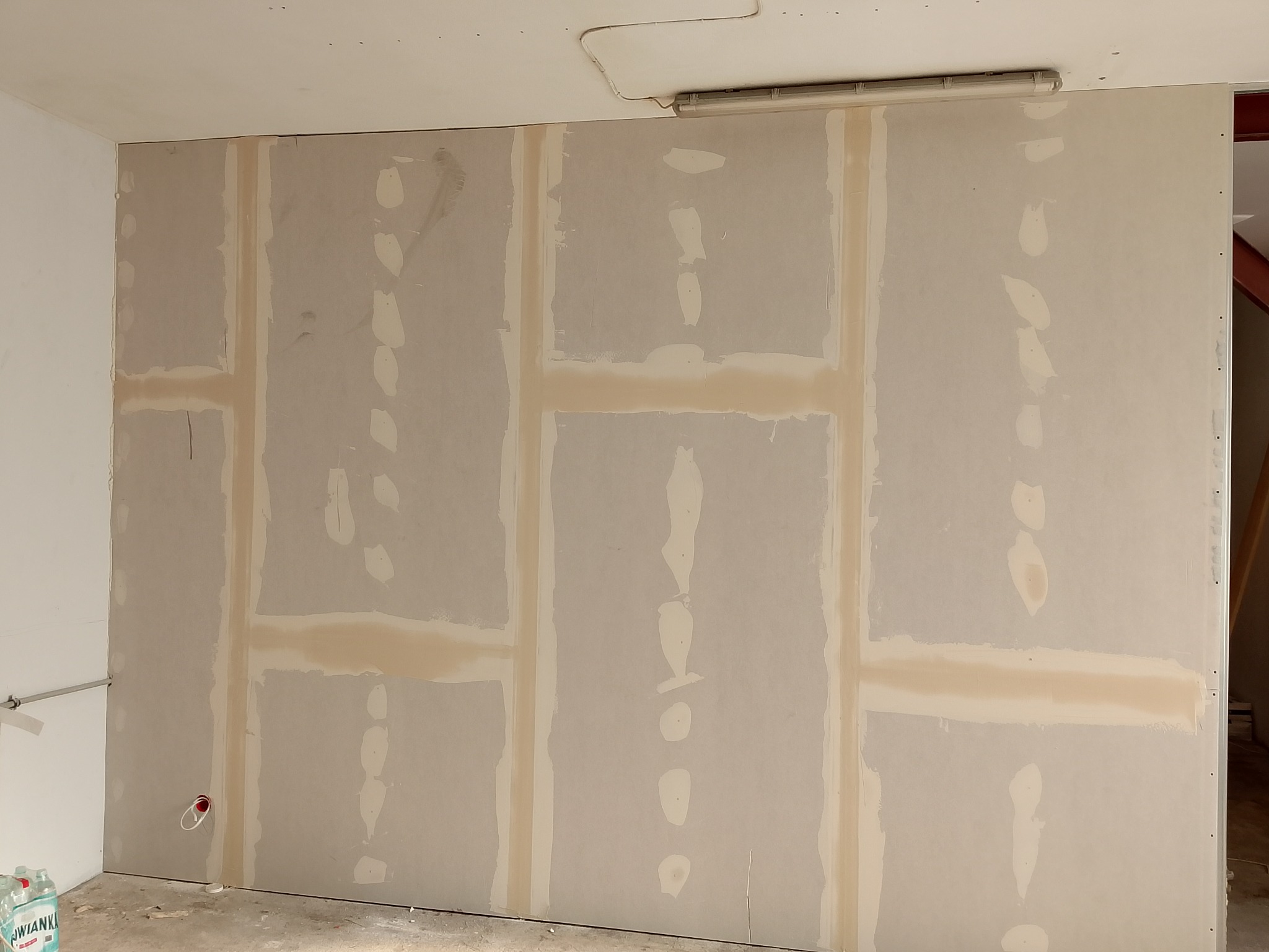 Ściana z płyt gipsowo-kartonowych, z widocznymi łączeniami i miejscami po szpachlowaniu. W tle widoczna lampa sufitowa i fragment konstrukcji dachu.