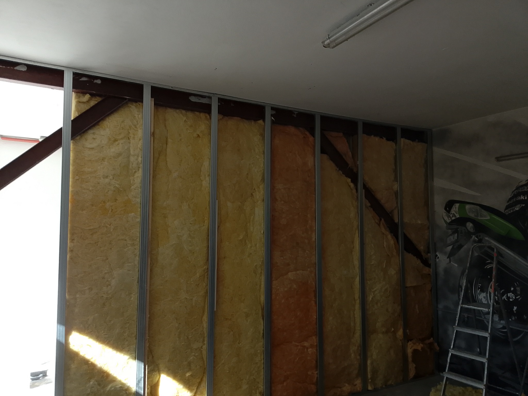 Ściana z izolacją z wełny mineralnej w metalowej konstrukcji, widoczne elementy mocujące. W tle malunek motocykla i drabina.