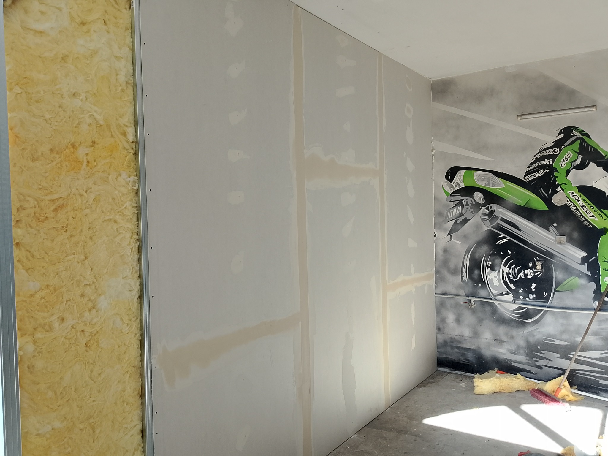 Wnętrze w trakcie prac: izolacja ścian wełną mineralną, płyty gipsowo-kartonowe z widocznymi łączeniami, na ścianie grafitowe graffiti motocykla.