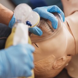Centrum Szkoleń Medycznych AQUAMED - Manekin do resuscytacji z założoną maską tlenową, ręce w niebieskich rękawiczkach, symulacja udzielania pierwszej pomocy.
