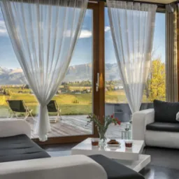 Jasny salon z widokiem na góry przez panoramiczne okna przesłonięte zwiewnymi, białymi firanami; na pierwszym planie białe meble wypoczynkowe i stolik z kwiatami oraz ciastkami.
