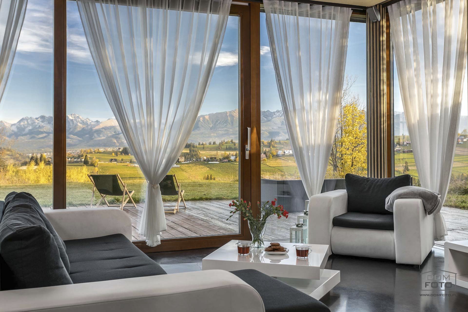Jasny salon z widokiem na góry przez panoramiczne okna przesłonięte zwiewnymi, białymi firanami; na pierwszym planie białe meble wypoczynkowe i stolik z kwiatami oraz ciastkami.