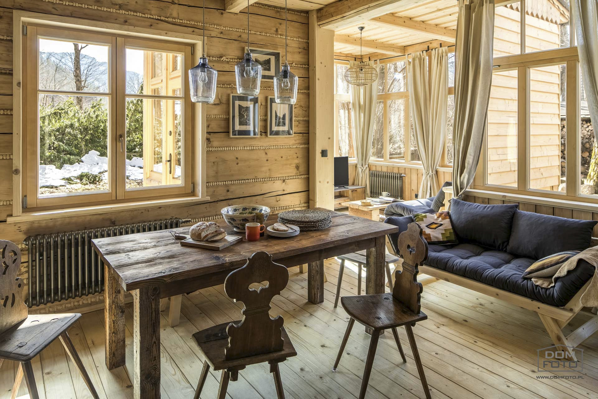 Rustykalne wnętrze domu z drewnianymi ścianami, stołem z zastawą, krzesłami z wyciętymi sercami i widokiem na ośnieżony ogród przez okno.