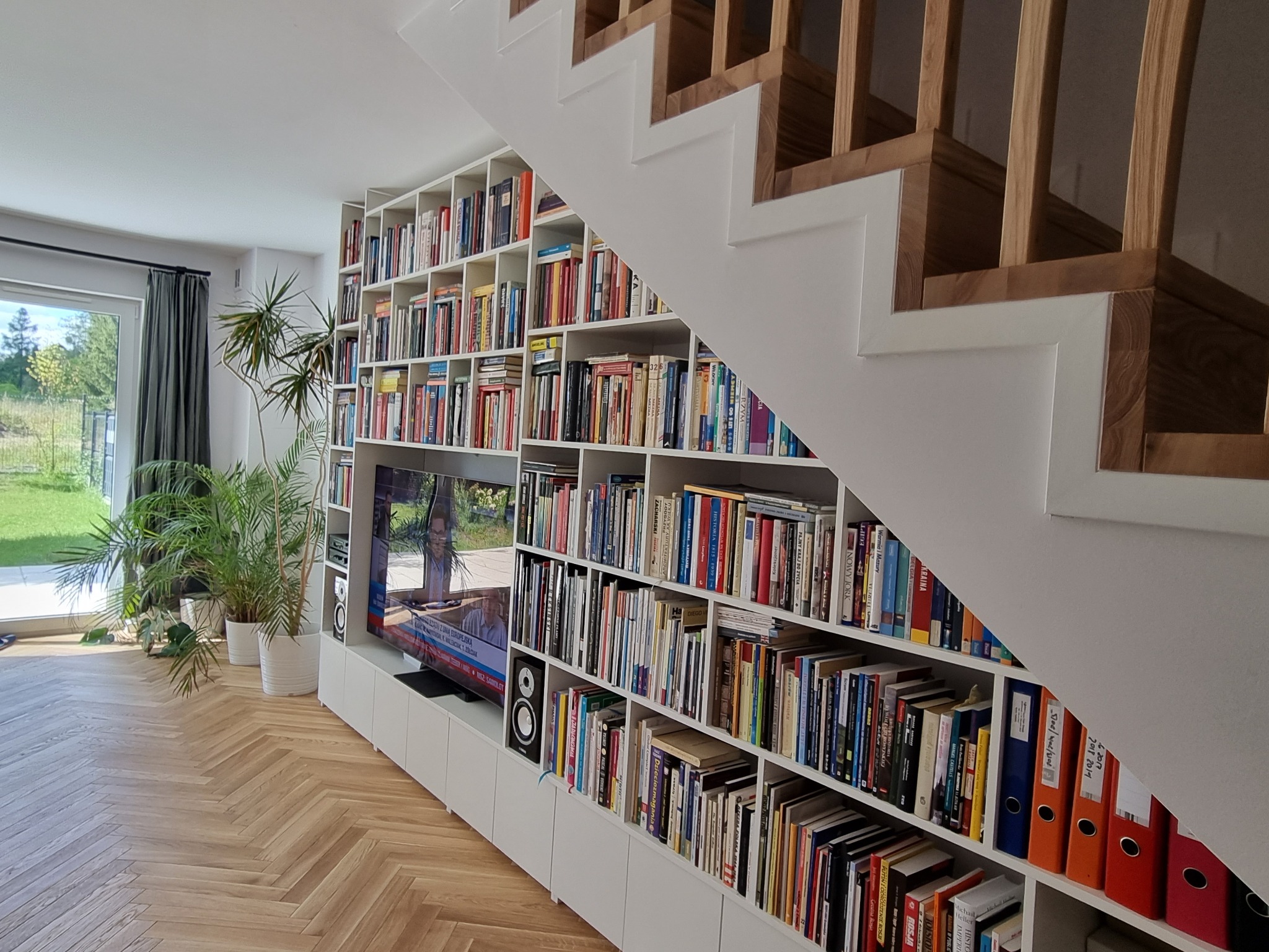 Salon z parkietem w jodełkę, zabudowaną biblioteczką pod schodami z telewizorem i roślinami doniczkowymi przy oknie.