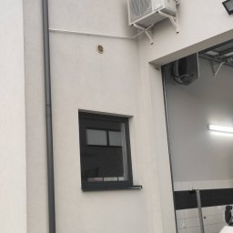 Ok Klima - Montaż i serwis klimatyzacji Kraków - Zewnętrzna jednostka klimatyzacji marki AUX zamontowana na białej ścianie budynku, widoczna rura odprowadzająca skropliny, obok okno i brama garażowa.