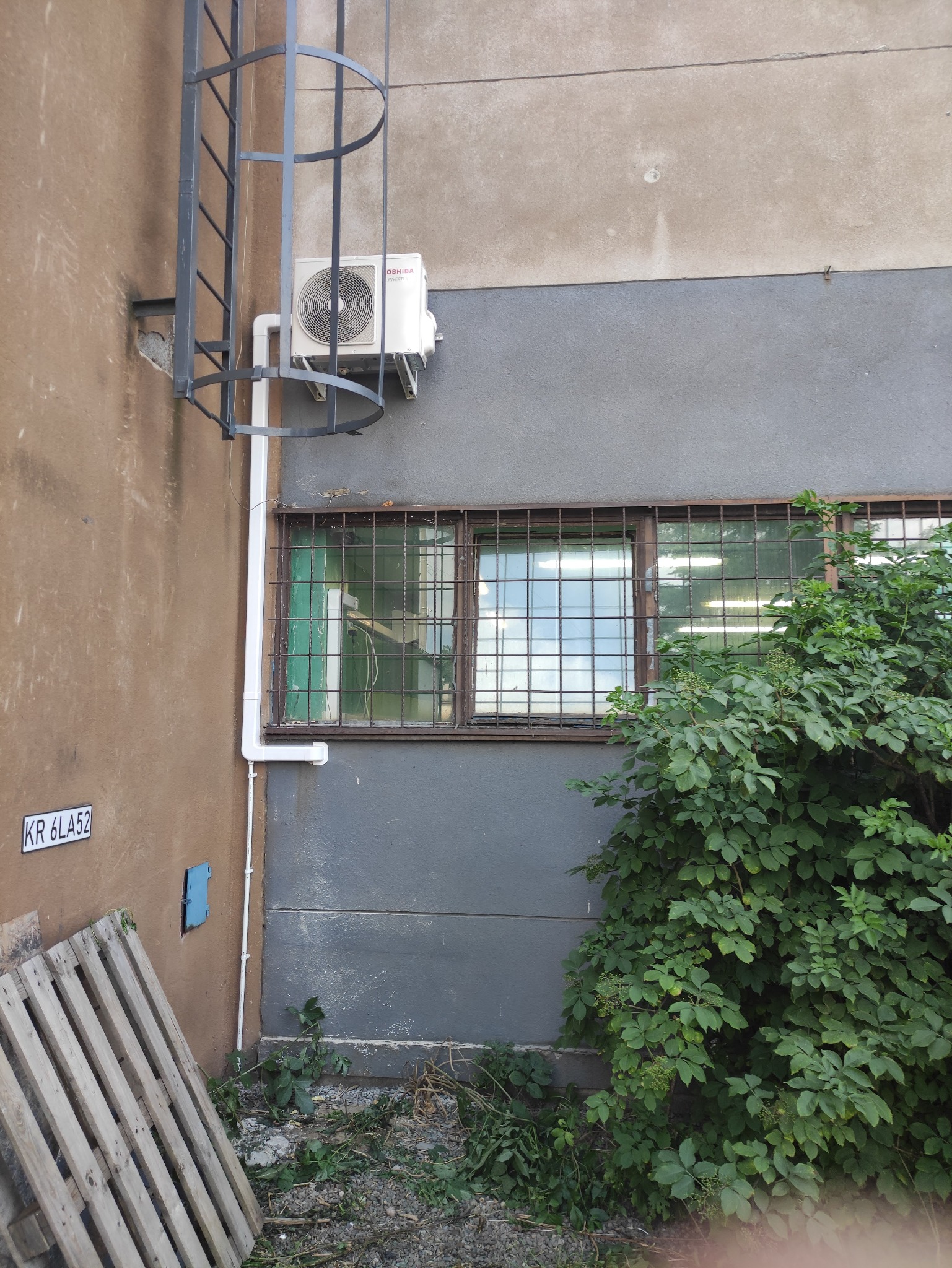 Zewnętrzna jednostka klimatyzacji Toshiba zamontowana na szarej ścianie budynku, obok metalowej drabiny i okna z kratami. Widoczne białe rury odprowadzające skropliny. Ziemia przy ścianie...