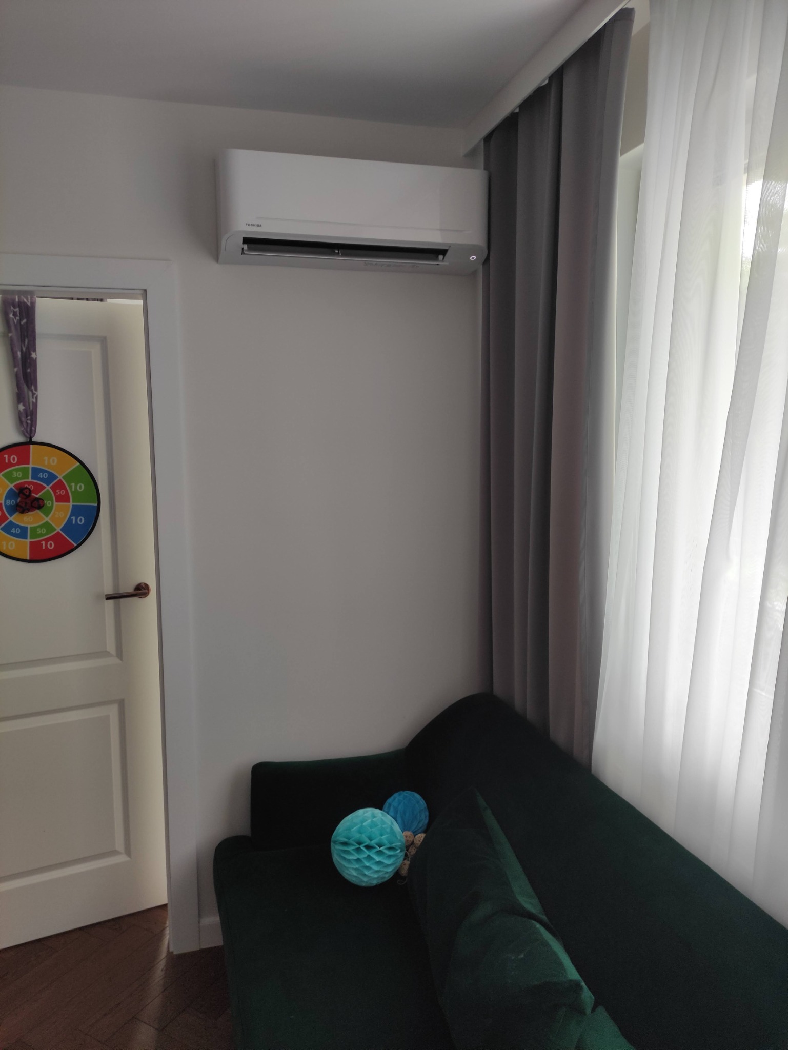 Biała klimatyzacja marki Toshiba zamontowana na ścianie w pokoju z zieloną sofą, obok zasłony w odcieniach szarości i białych firanek, widoczny cel do darta na drzwiach.