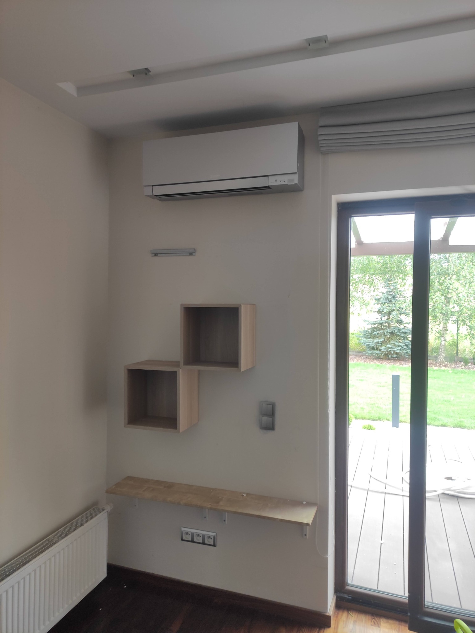 Zamontowana jednostka klimatyzacji w domu w Krakowie, widoczna nad drewnianymi półkami ściennymi i grzejnikiem, z oknem balkonowym w tle.