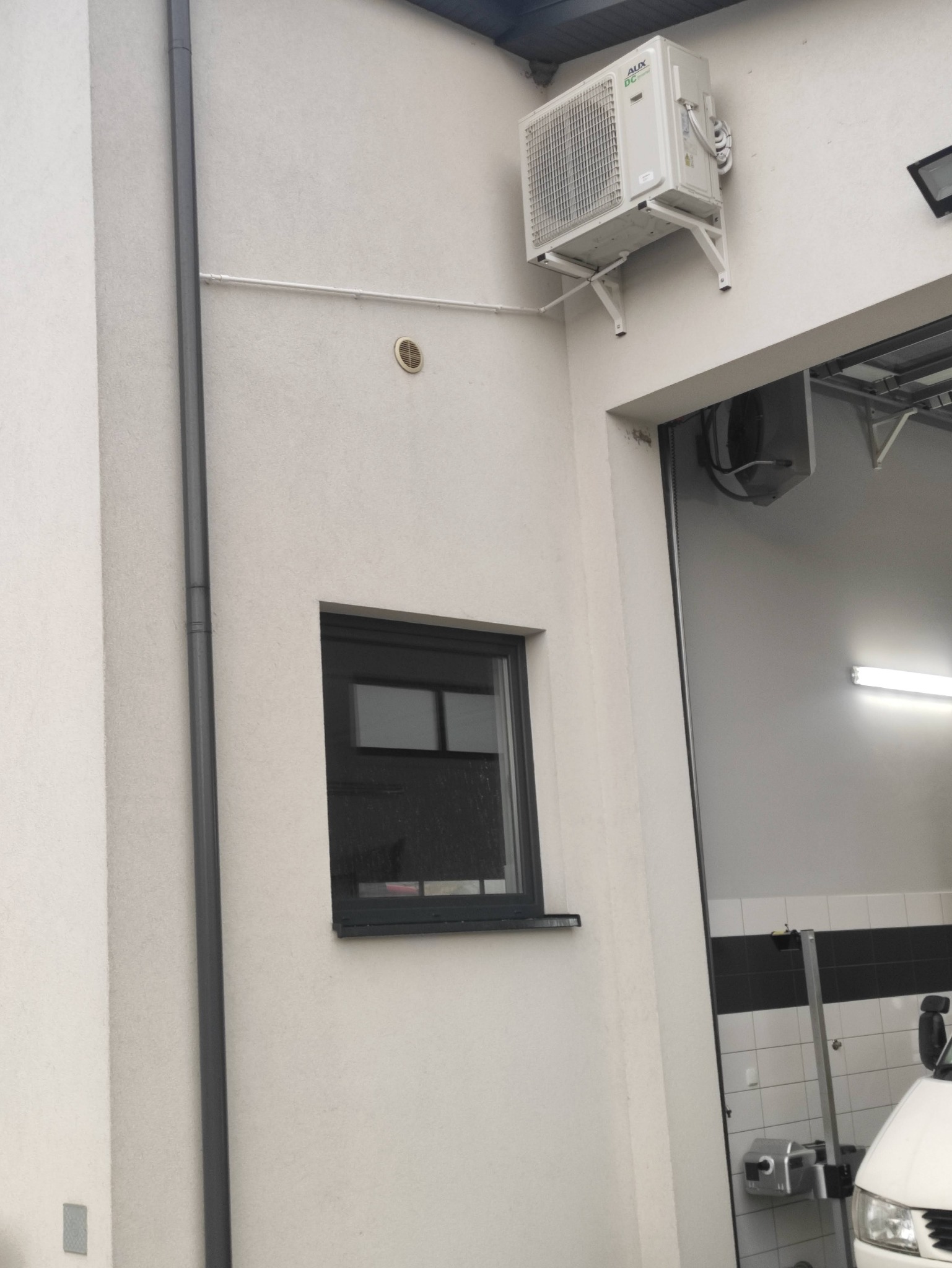 Zewnętrzna jednostka klimatyzacji marki AUX zamontowana na białej ścianie budynku, widoczna rura odprowadzająca skropliny, obok okno i brama garażowa.